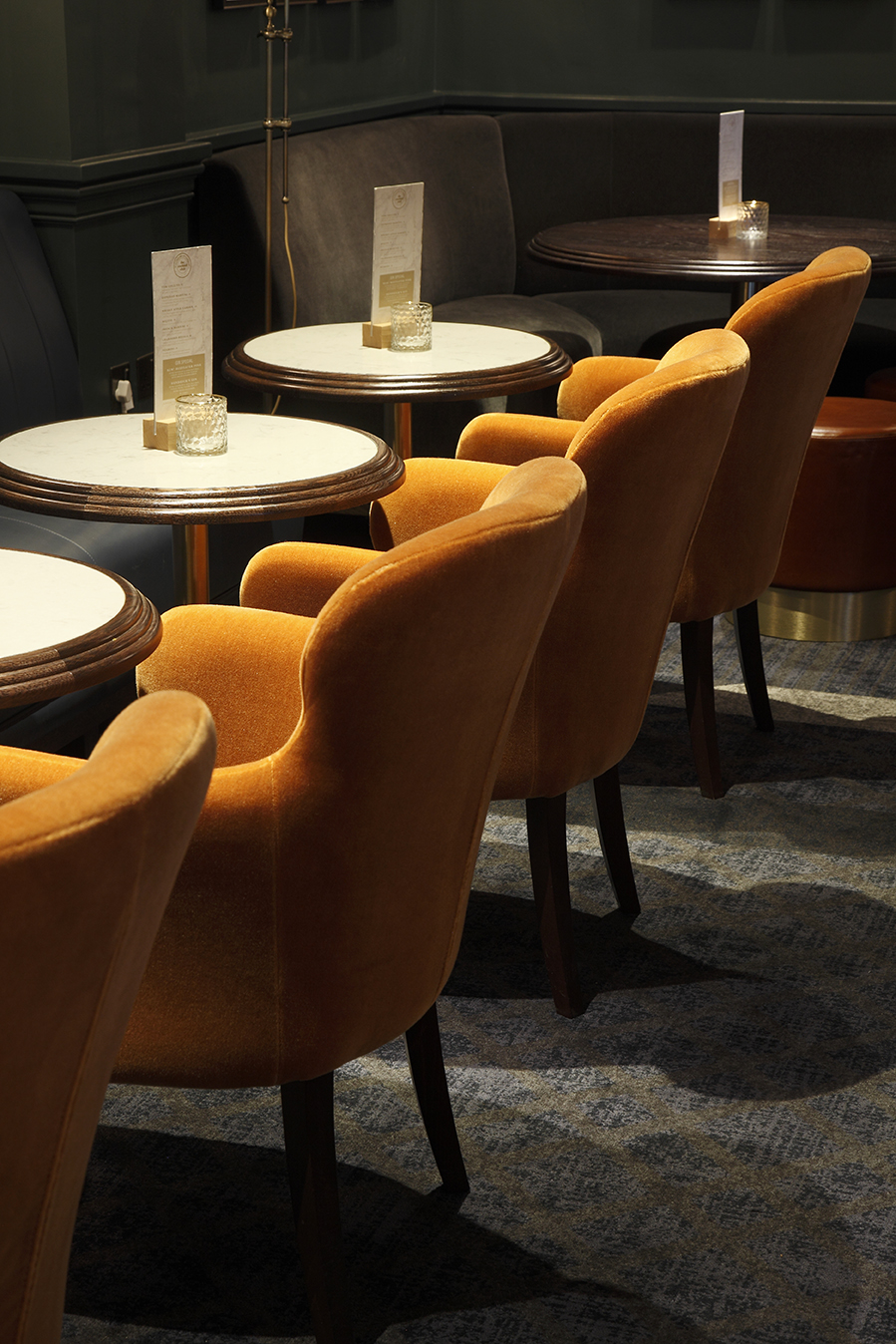 A row of orange velvet armchairs and circular white topped tables with menus on in a bar with moody lighting
