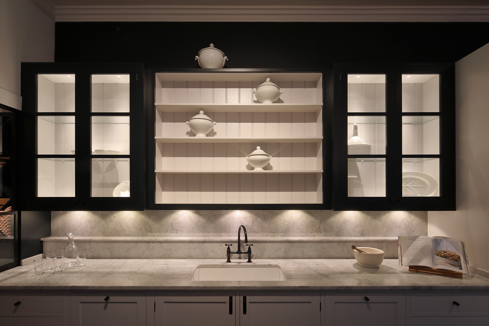 Show kitchen with open shelves and glazed cabinetry above a marble worktop and  basin, with dramatic lighting