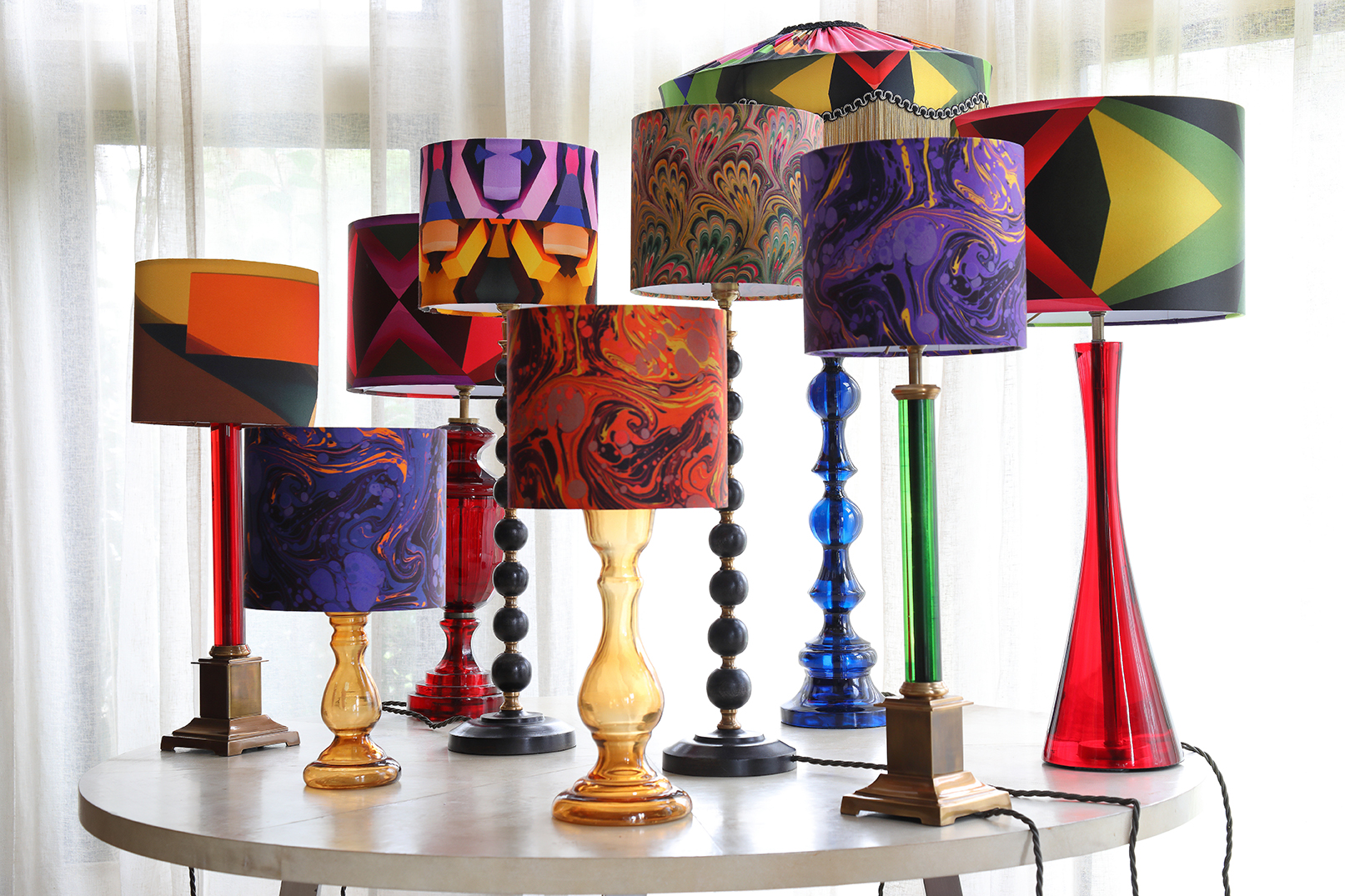A collection of lamps with colourful patterned shades and coloured glass stands on a circular table in front of windows.
