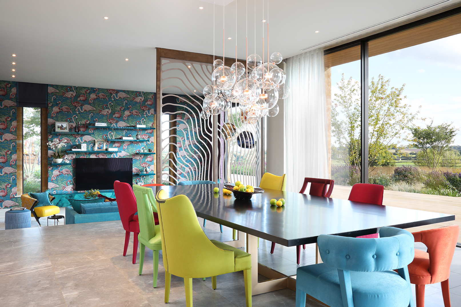 Open plan dining area with sunken seating area beyond a wavy metal screen. Mix and match coloured upholstered chairs around the table.