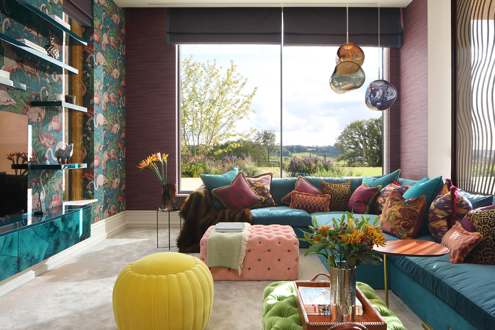 Colourful living room with huge windows framing views across the Cotswolds. Flamingo wallpaper and a teal coloured corner sofa with numerous cushions and button backed ottomans.