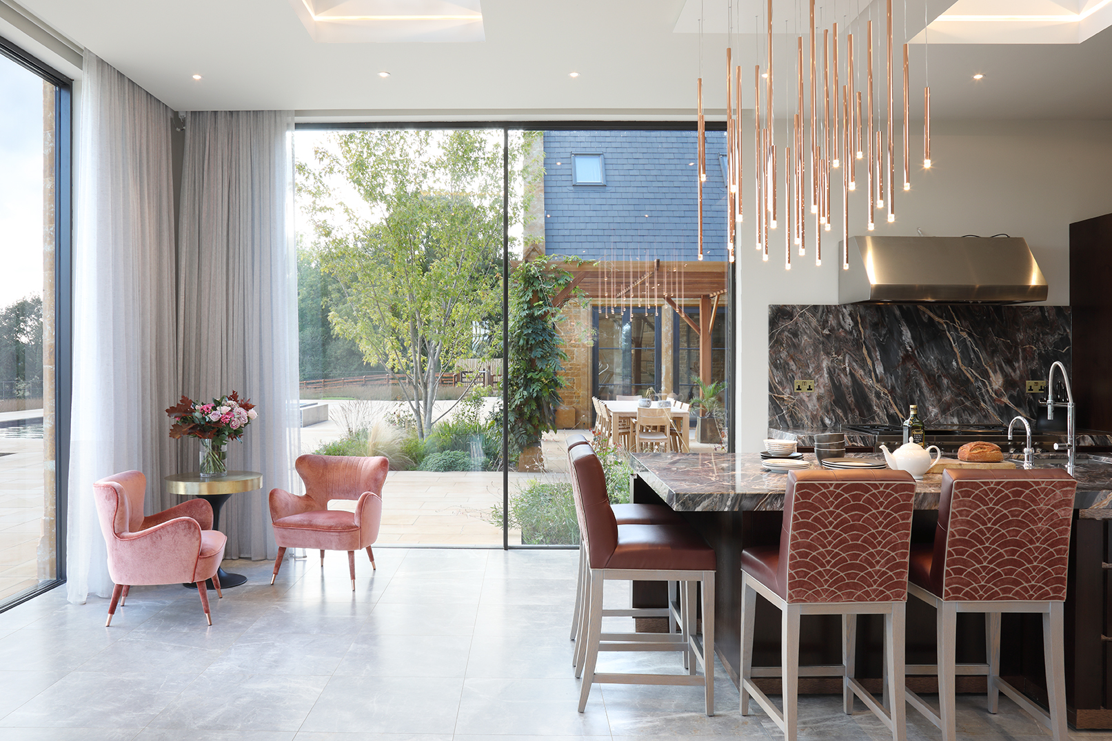Brown marble kitchen with terracotta coloured upholstered barstools at central island. Full height sliding glass doors framed by sheer curtains frame the garden beyond.