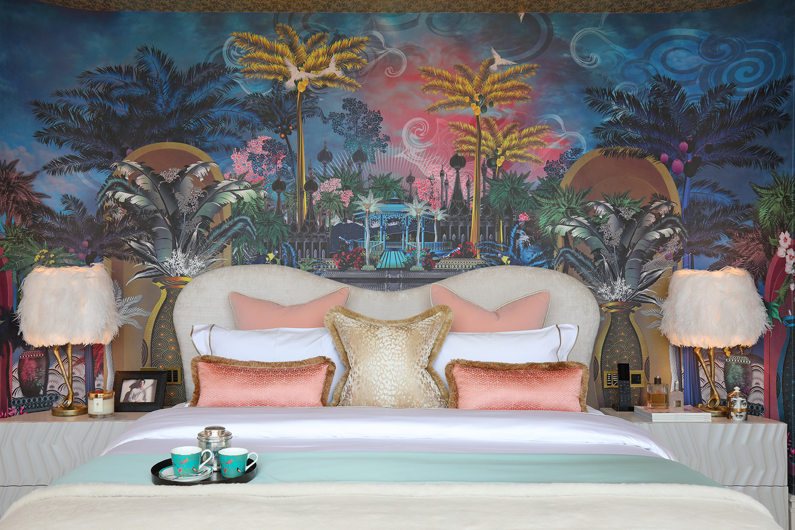 Opulent master bedroom with wallpaper fantasy panorama with palm trees and exotic architecture. A tray with coffee cups sits on the bed, and fluffy bird like lamps are on the bedside tables.