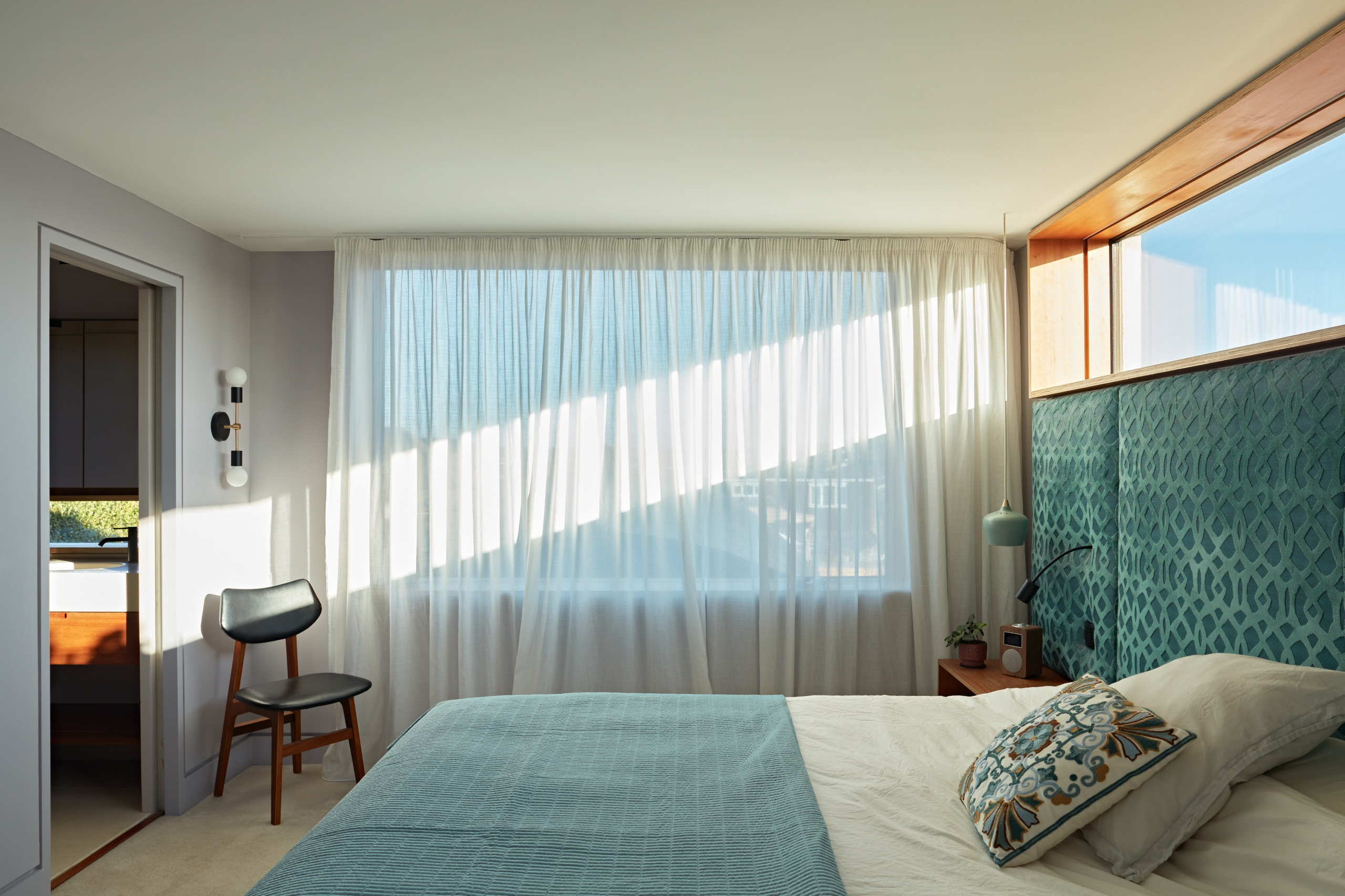 A hotel inspired bedroom with sheer drapes and rich velvet fabrics on a bespoke full width headboard. The high level timber framed window adds light and an unexpected sense of drama.