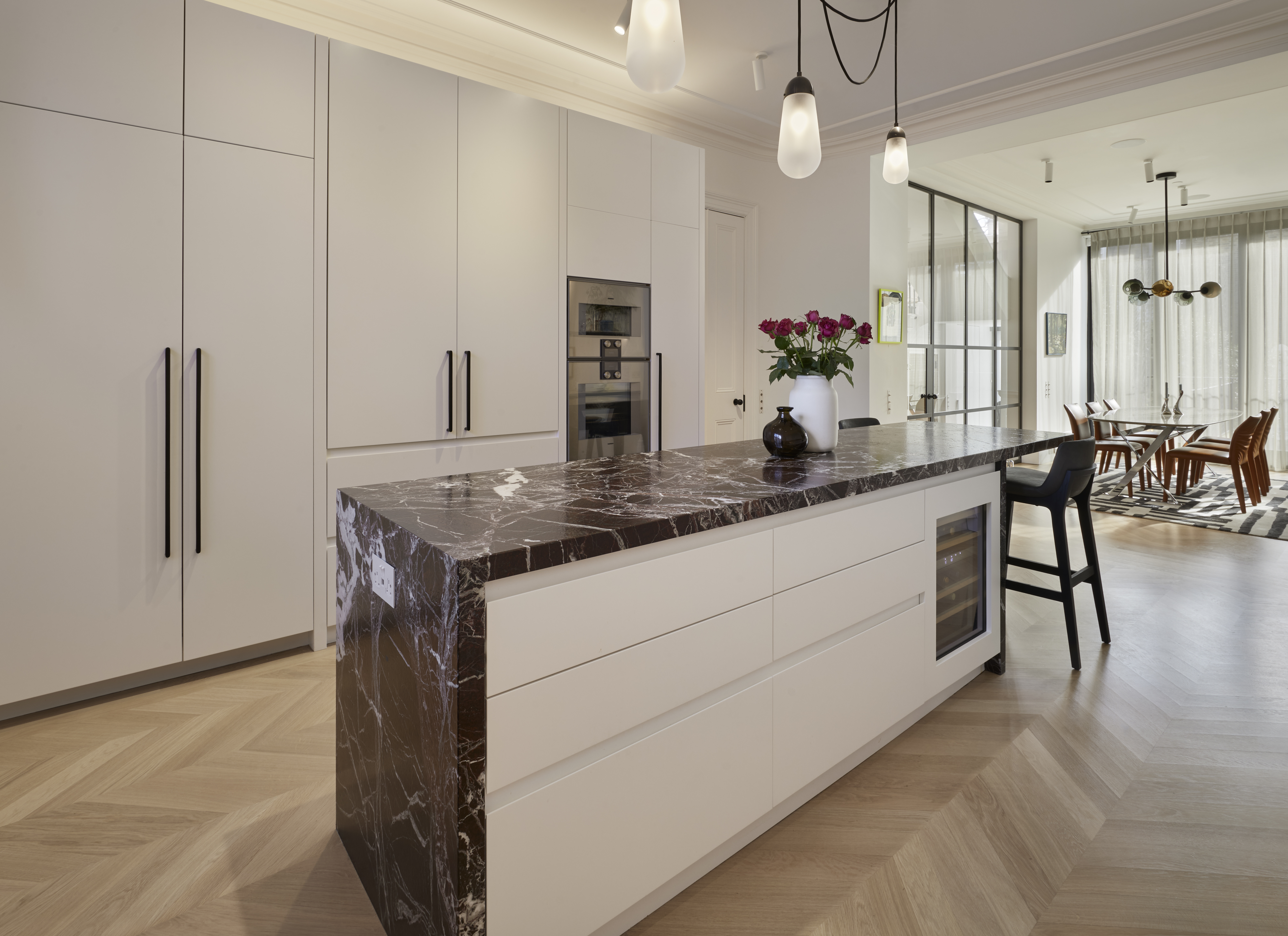 View of the front of a white painted kitchen island with a marble worktop & sides & tall cabinets on teh opposite wall.