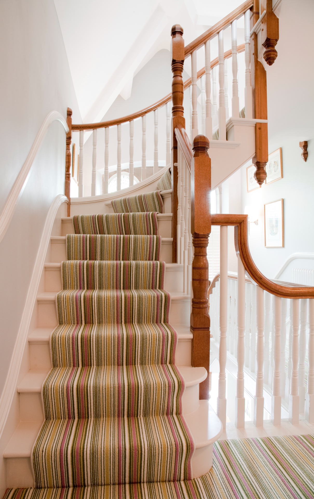 A Victorian style staircase with elaborate finials & wooden handrail with a multishade green striped carpet runner.