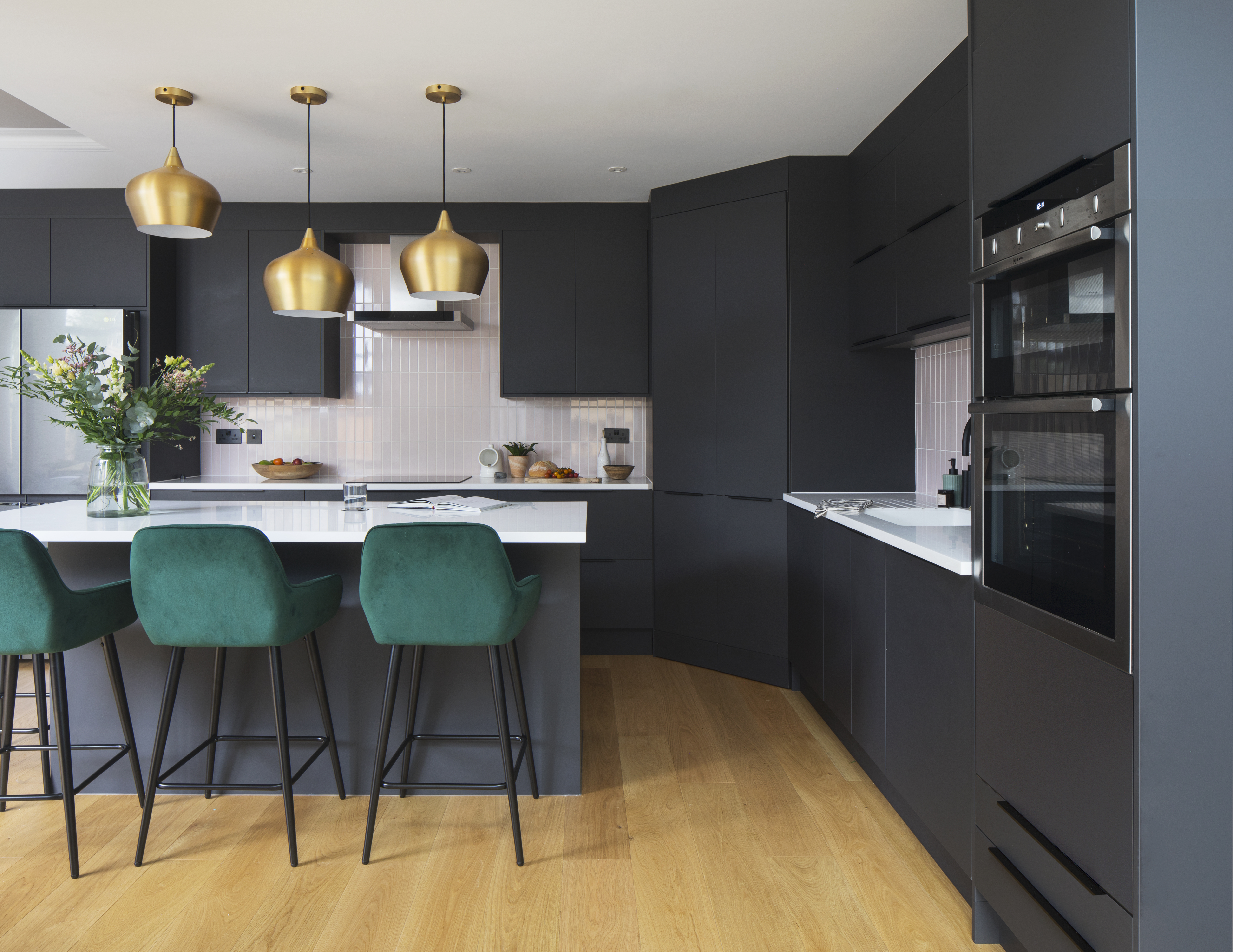 Black kitchen units with white solid surface worktop.  Kitchen island with green velvet bar stools.  Brass industrial style pendant lights over island