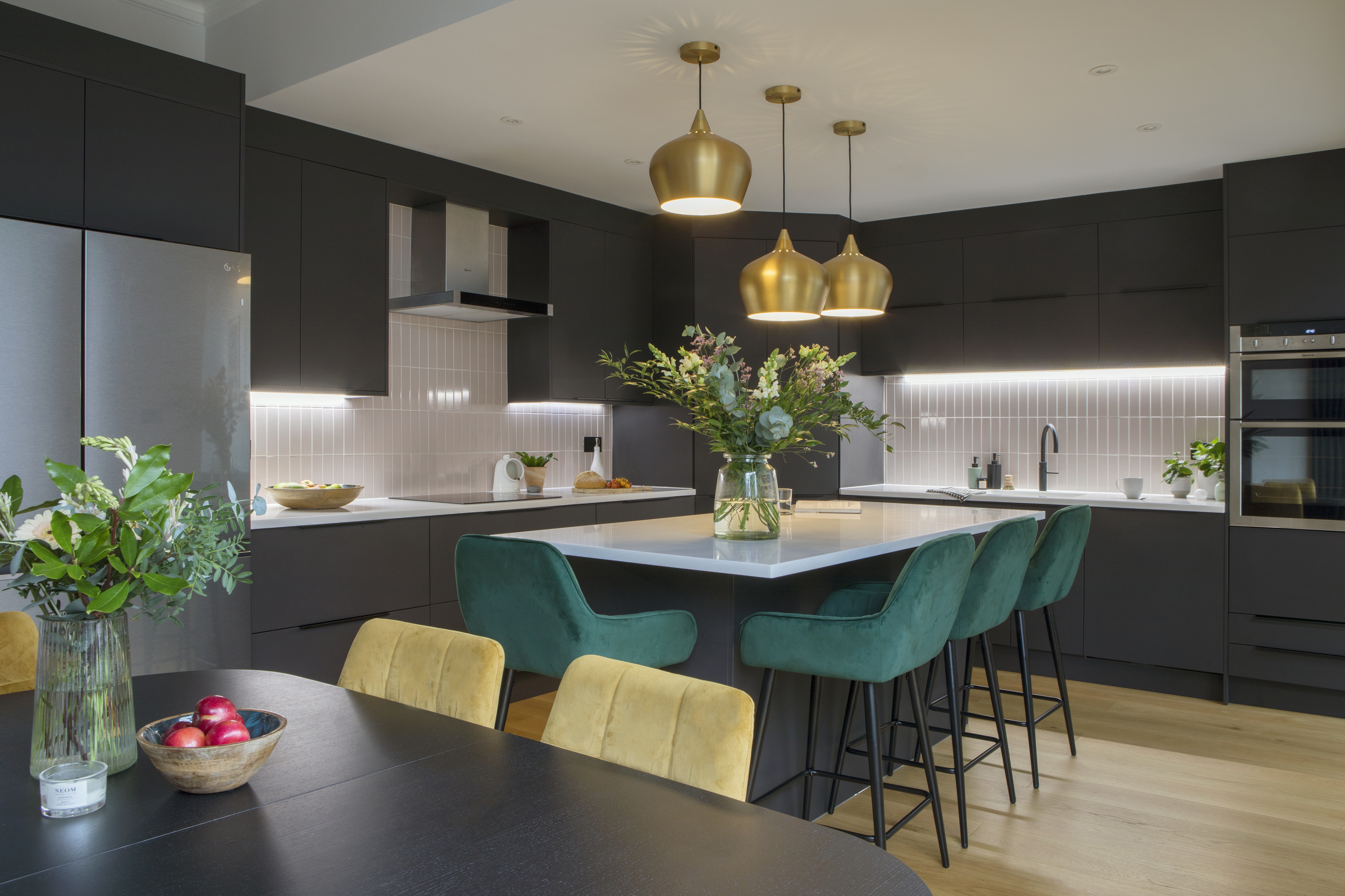 Black kitchen units with white solid surface worktop.  Kitchen island with green velvet bar stools.  Brass industrial style pendant lights over island.  Black dining table and mustard chairs