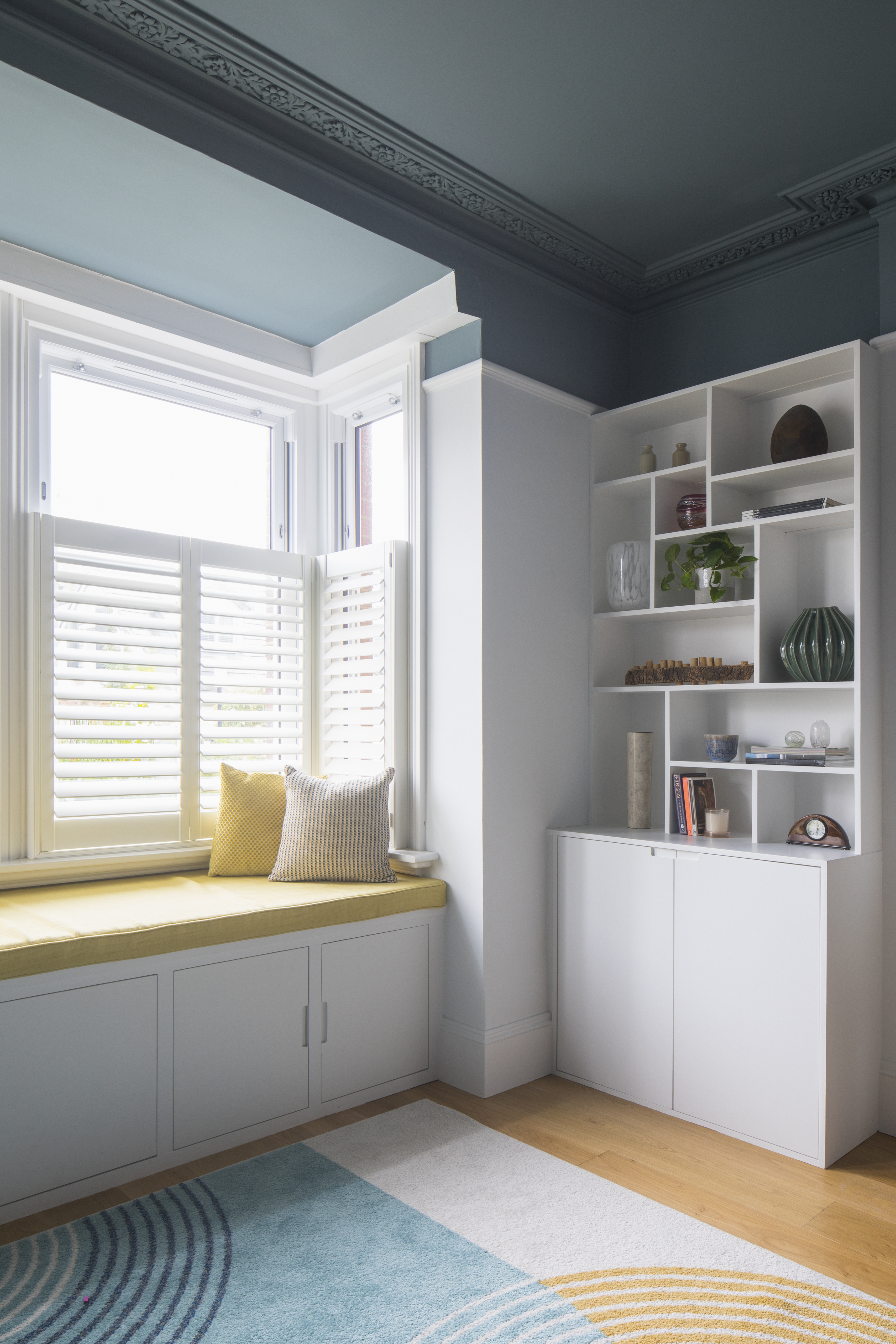 Window seat with mustard cushion.  Window shutters.  Mid century shelf and cupboard alcove storage.  Dark blue ceiling. 