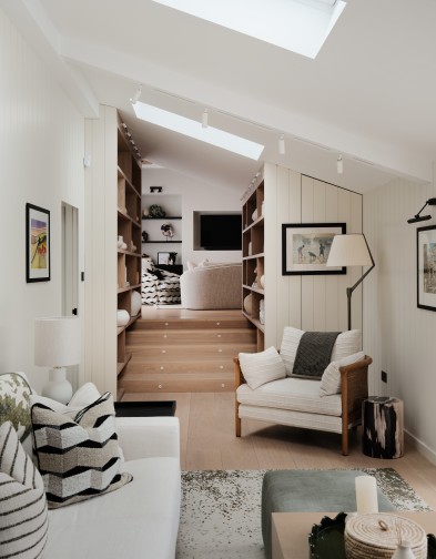Narrow room with stairs leading to another lounge area. Cream sofa and armchair with rattan back. Staircase running either side of the staircase. Tongue and groove panelling all over.