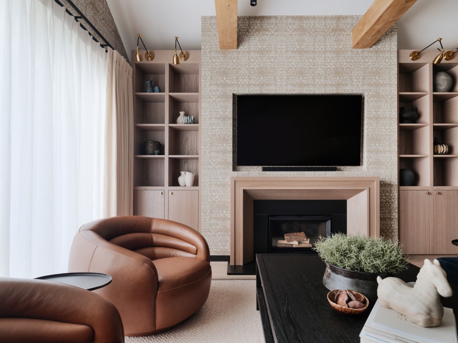 Lounge with brown leather armchairs sat in front of fireplace with wooden fire surround and matching wooden joinery either side. Feature lighting attached to the joinery and leaning over to light each nook.
