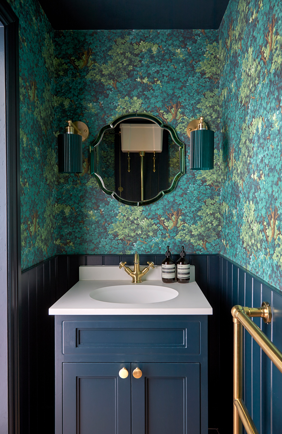 A small cloakroom with rich teal and navy tones. The walls feature a leafy patterned wallpaper with dark panelling below, complemented by brass fixtures and lighting. A scalloped mirror and navy vanity with a white countertop complete the bold, elegant look. A small cloakroom with rich teal and navy tones. The walls feature a leafy patterned wallpaper with dark panelling below, complemented by brass fixtures and lighting. A scalloped mirror and navy vanity with a white countertop complete the bold, elegant look.