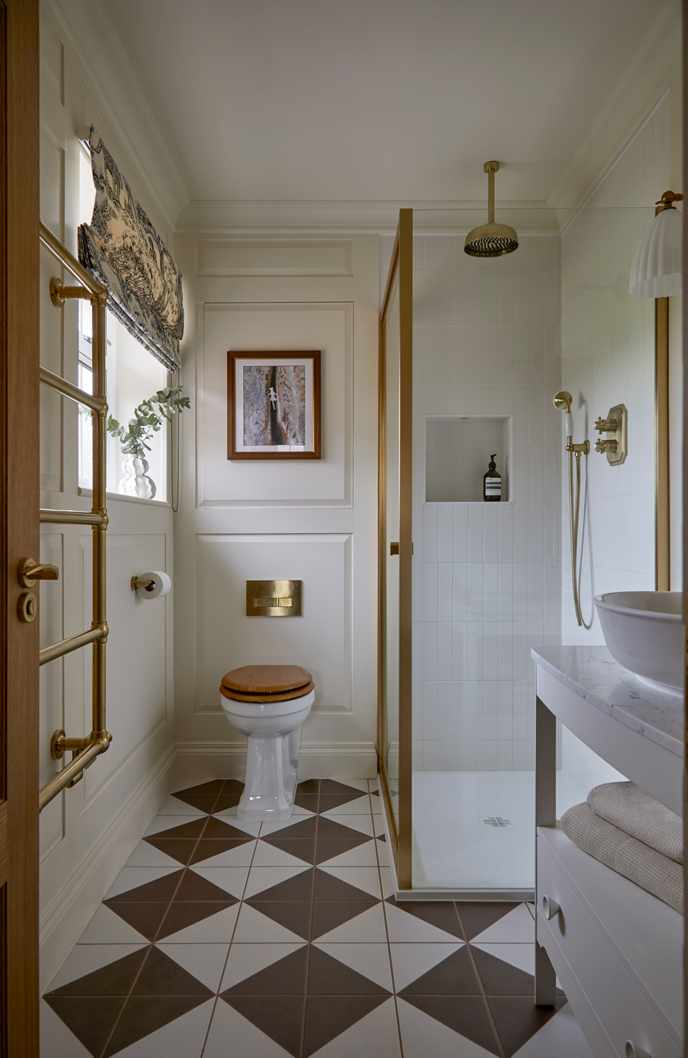 A refined shower room with cream panelling, brass fittings, and geometric floor tiles in brown and white. It features a glass shower enclosure, a wooden toilet seat, and a marble-topped vanity, with a patterned Roman blind adding a touch of elegance. A refined shower room with cream panelling, brass fittings, and geometric floor tiles in brown and white. It features a glass shower enclosure, a wooden toilet seat, and a marble-topped vanity, with a patterned Roman blind adding a touch of elegance.