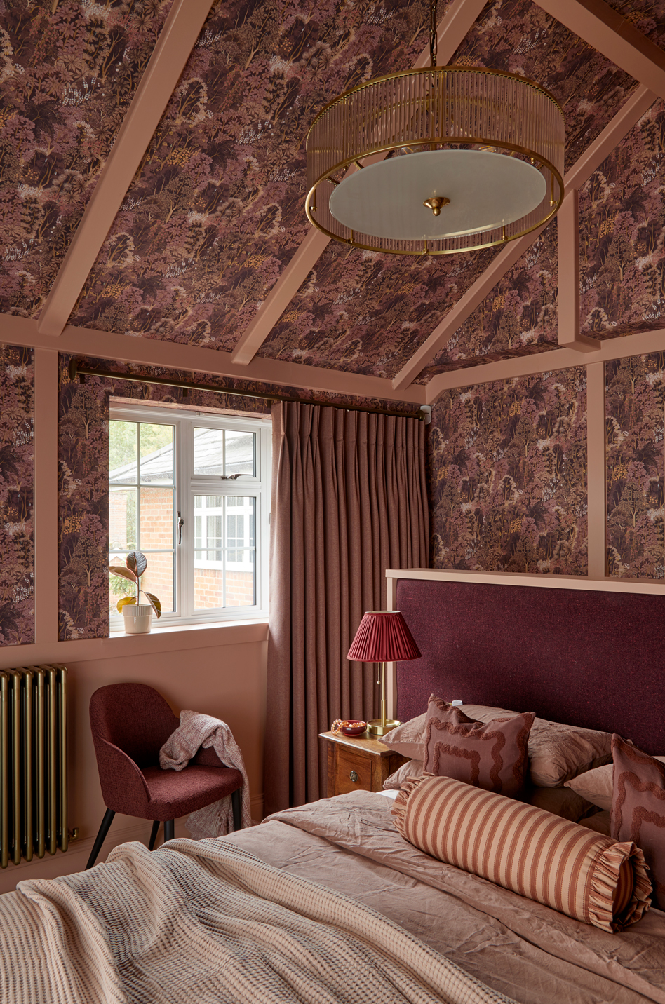 A cosy bedroom in deep pink and plum tones with patterned wallpaper across walls and ceiling. The bed has layered linens and cushions in warm shades, with a brass lamp, soft curtains, and a vintage-style pendant light adding warmth and texture. A cosy bedroom in deep pink and plum tones with patterned wallpaper across walls and ceiling. The bed has layered linens and cushions in warm shades, with a brass lamp, soft curtains, and a vintage-style pendant light adding warmth and texture.