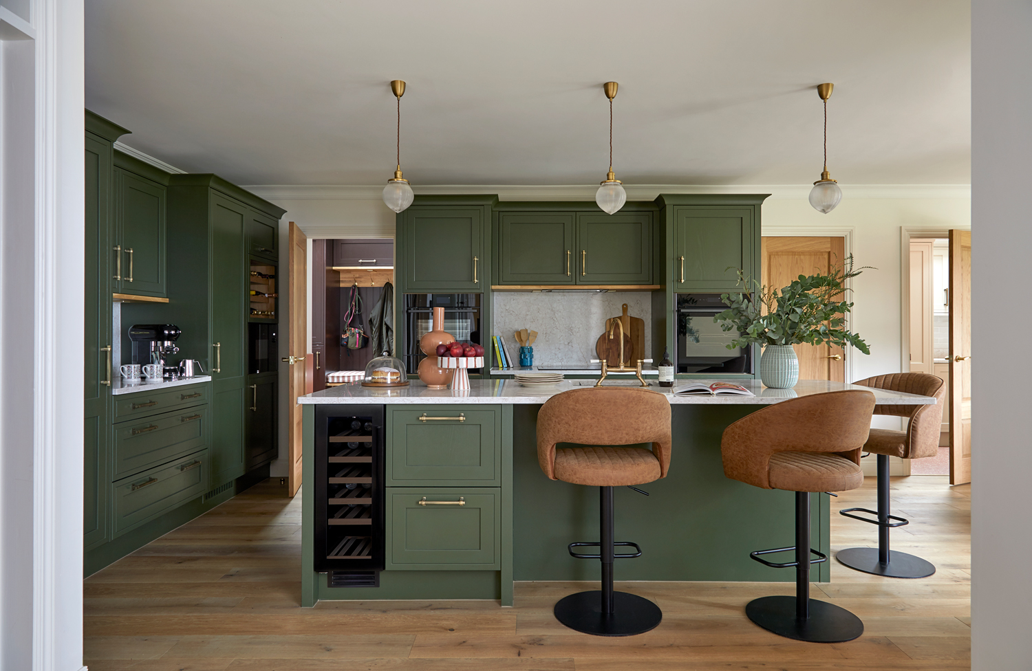 A welcoming kitchen with olive green cabinetry, brass hardware, and warm oak flooring. A marble-topped island with tan bar stools sits at the centre, complemented by globe pendant lights and soft brass accents that add warmth and balance. A welcoming kitchen with olive green cabinetry, brass hardware, and warm oak flooring. A marble-topped island with tan bar stools sits at the centre, complemented by globe pendant lights and soft brass accents that add warmth and balance.