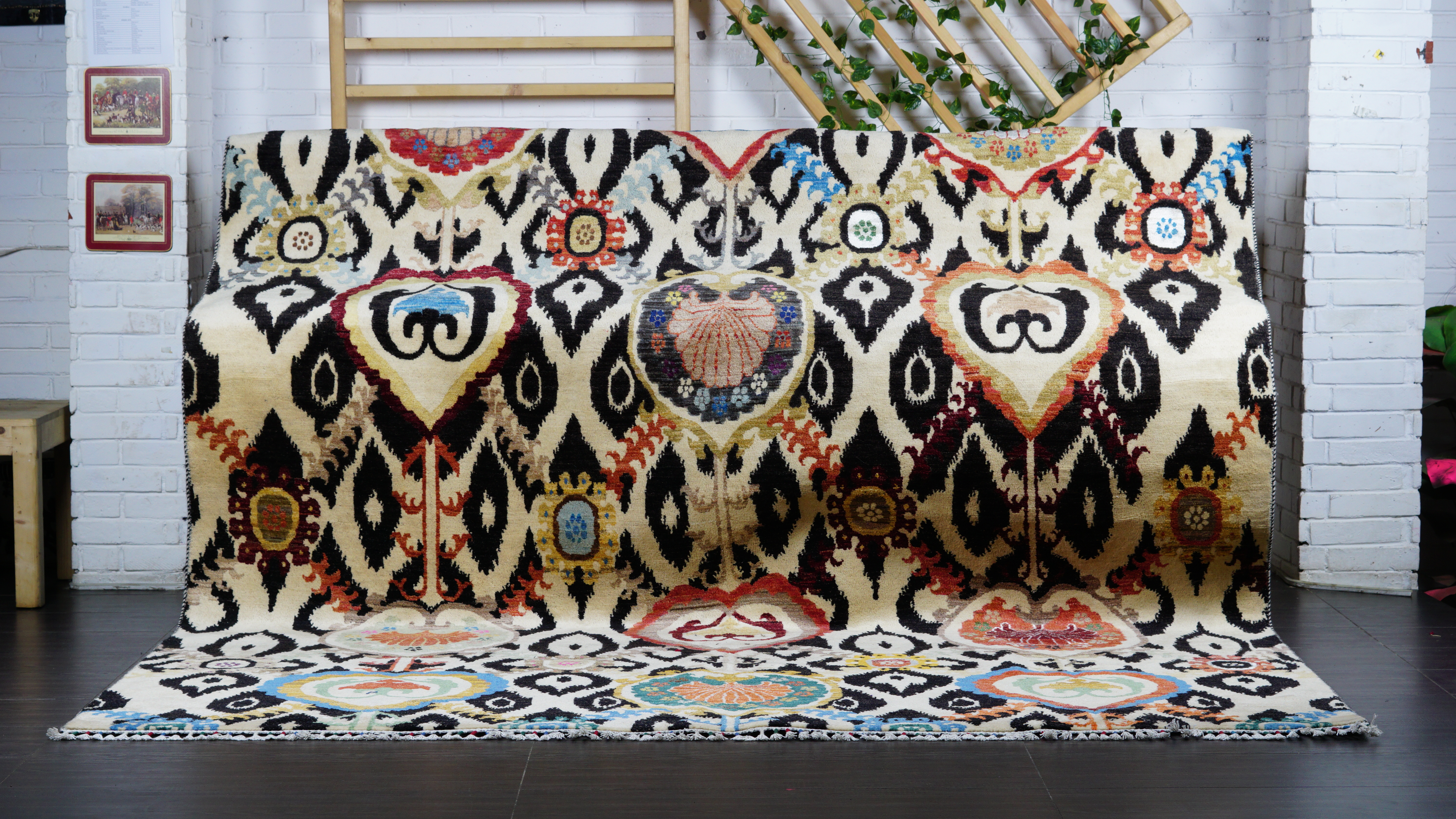 A large, hand-knotted wool and silk rug with a bold, symmetrical, modern Ikat pattern in black, red, and blue on a cream background, draped over a sofa in a studio.