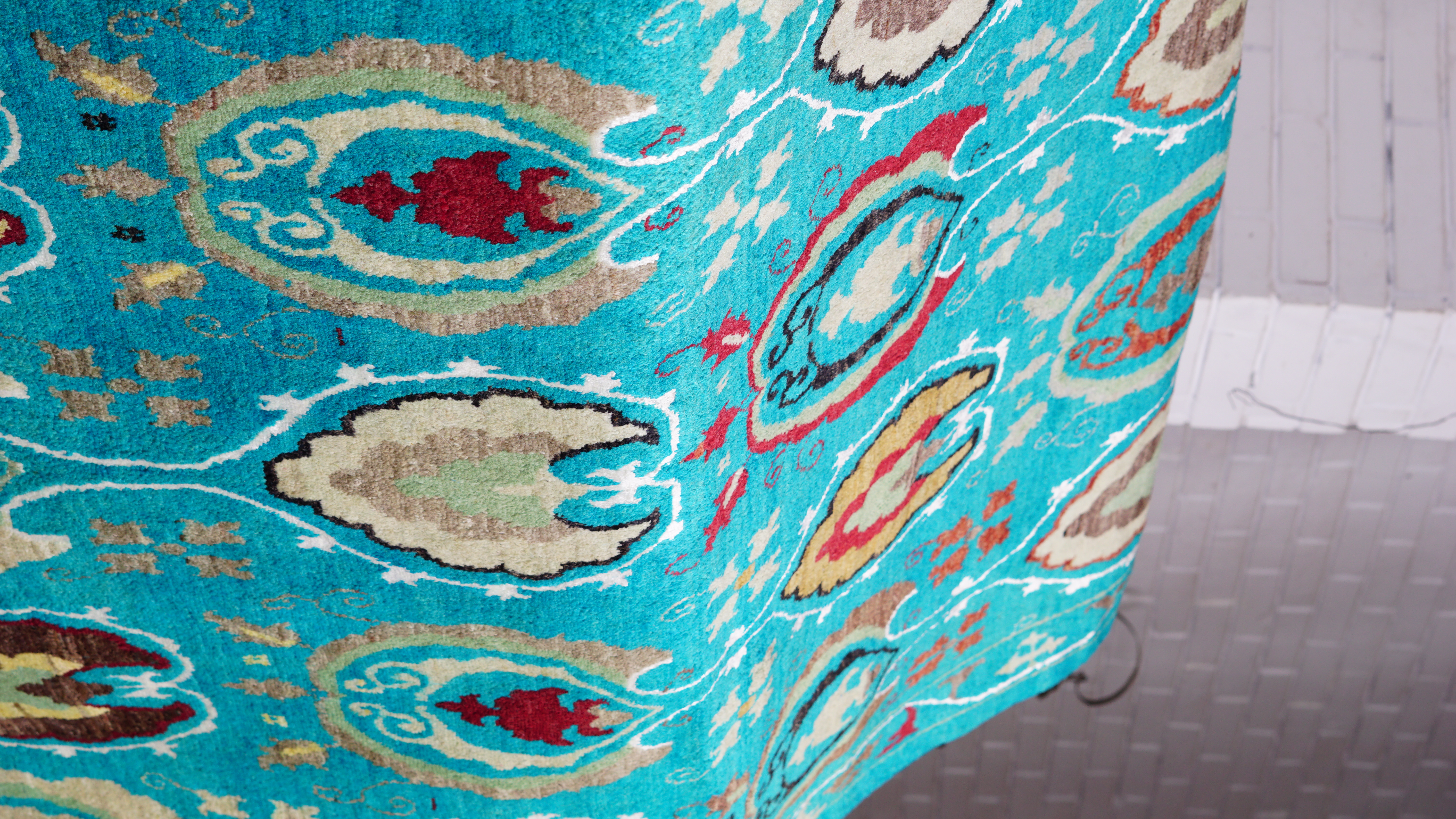 A close-up, angled view of a hand-knotted wool and silk rug in a vibrant turquoise, featuring a transitional pattern with red, beige, and brown motifs, draped over a surface.