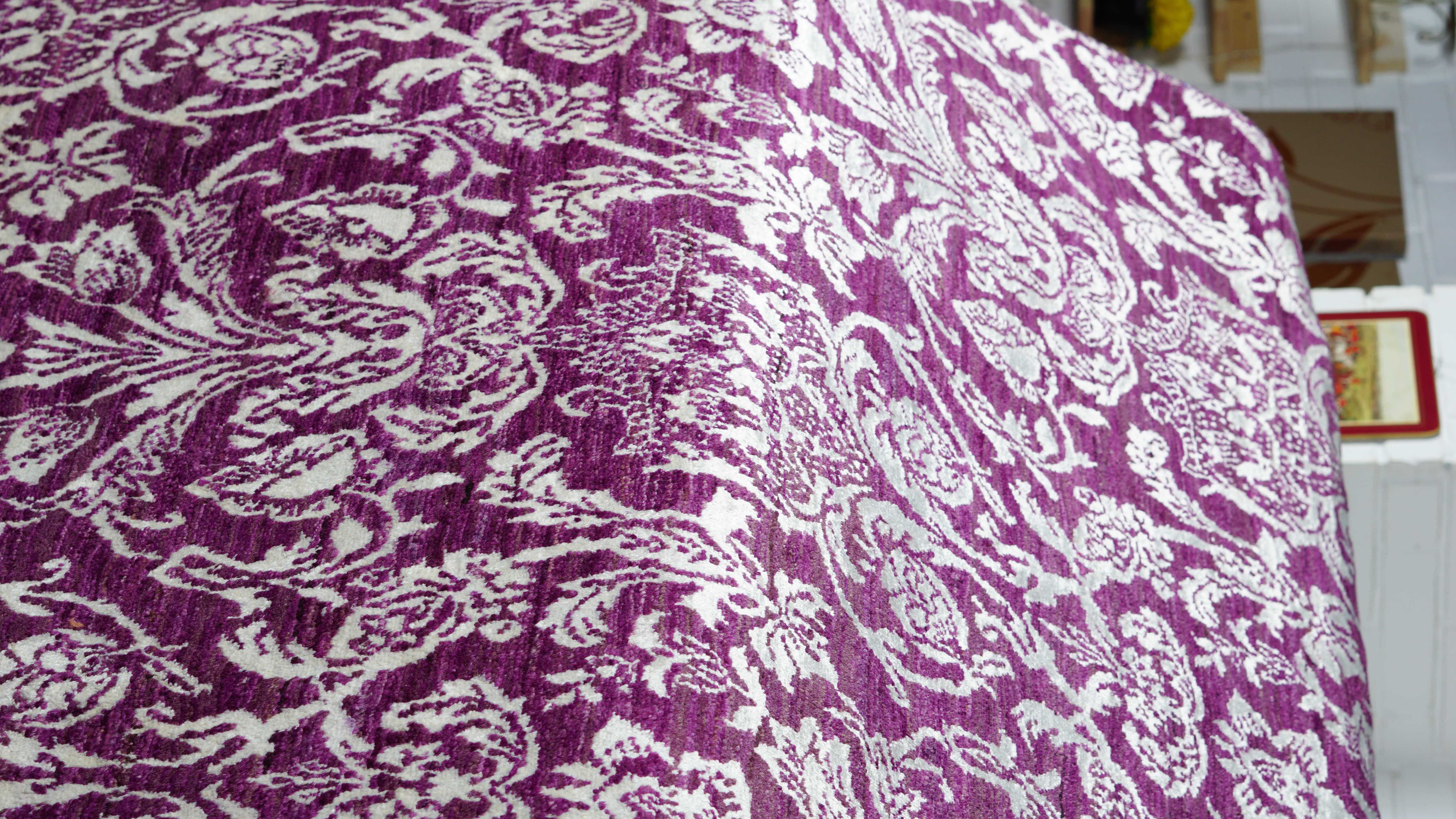 A close-up, angled view of a hand-knotted wool and silk rug featuring an intricate, white floral damask pattern on a deep purple background.