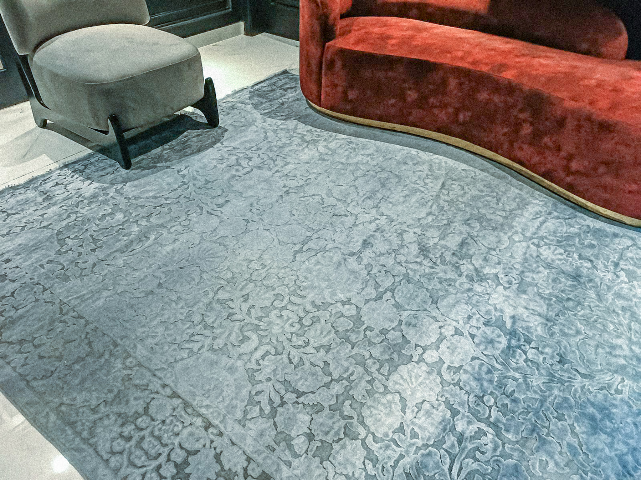 A close-up, angled view of a luxurious, light-blue silk-wash rug featuring a subtle, tonal floral pattern, with a red velvet chaise lounge partially visible.