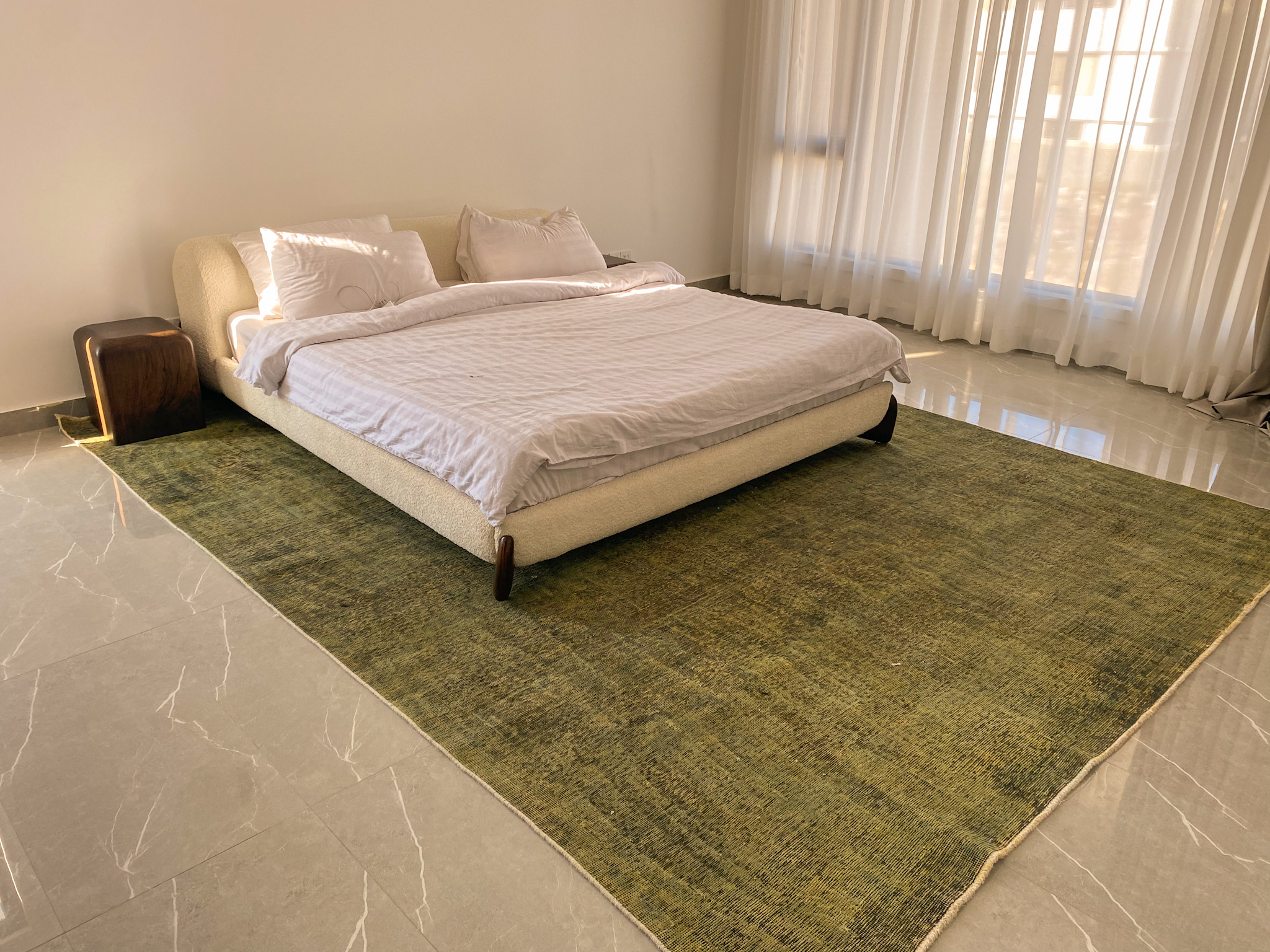 A large, overdyed Persian rug in a distressed, olive-green tone, placed under a modern, cream-coloured bed in a minimalist bedroom with grey tile floors.