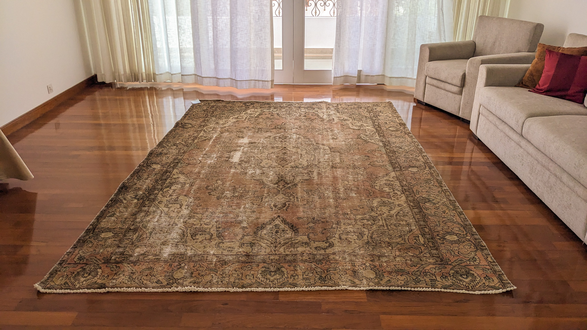 A large, overdyed Persian rug with a distressed, vintage look, in a faded, earthy brown and terracotta tone, placed on a polished hardwood floor in a living room.