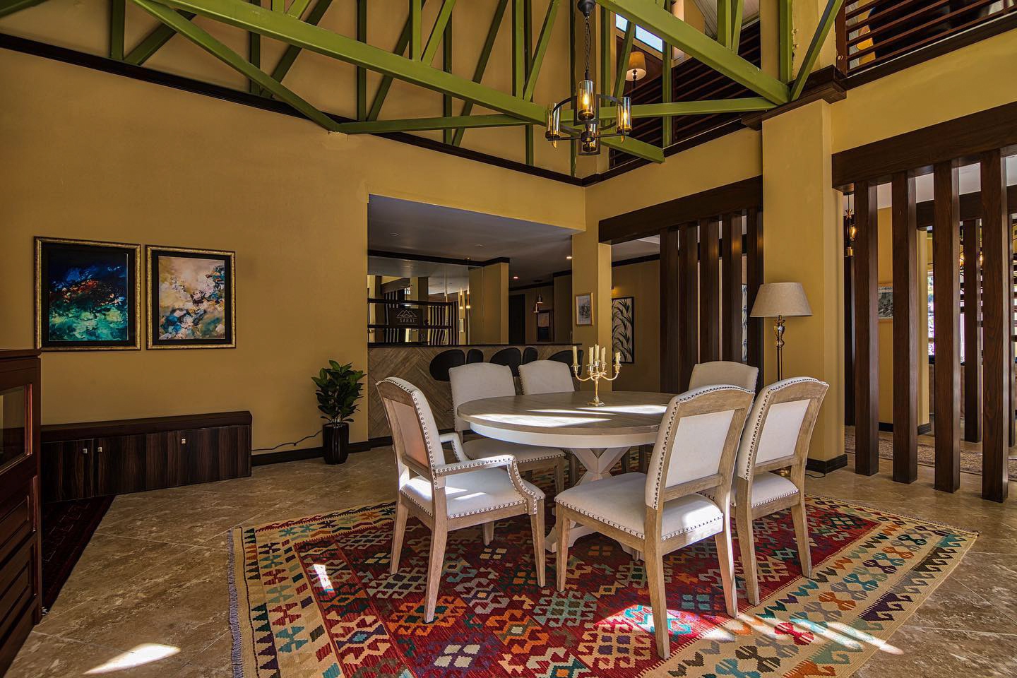A dining room featuring a large, geometric kilim rug in red and orange under a round white wooden table with six chairs. The room has yellow walls and an exposed green-painted-truss ceiling.