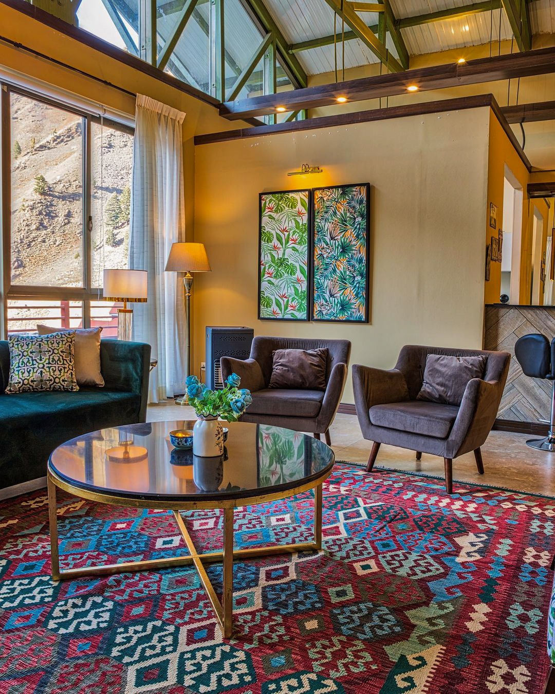 A lounge area in a mountain retreat, featuring a large, geometric kilim rug in red and blue, a dark green sofa, two brown armchairs, and a round black coffee table.