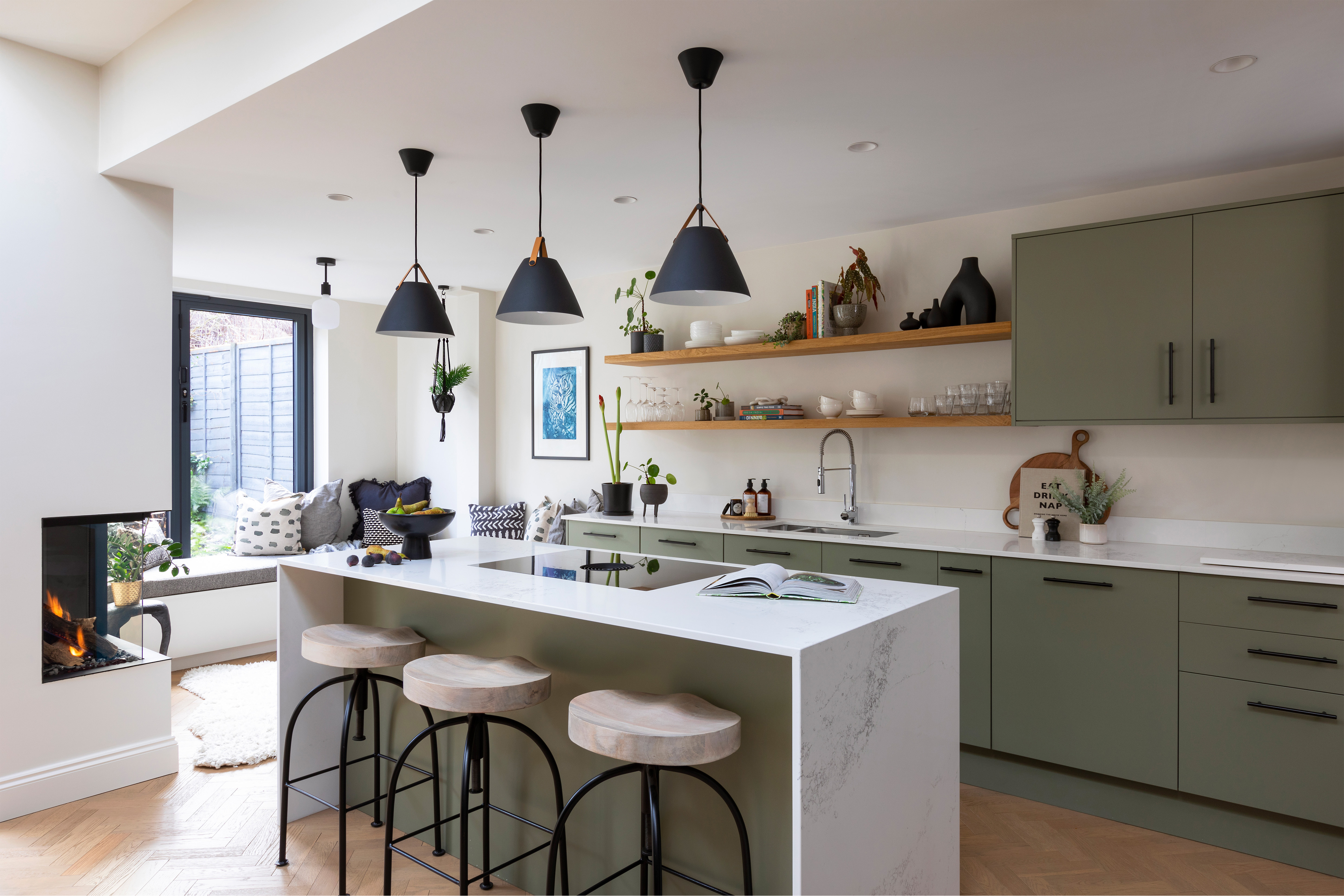 Modern open-plan kitchen with sage-green cabinetry, white worktops, timber open shelving, black pendant lights, and a central island with three wooden stools. A window seat, indoor plants, artwork and a contemporary fireplace add warmth and texture.
