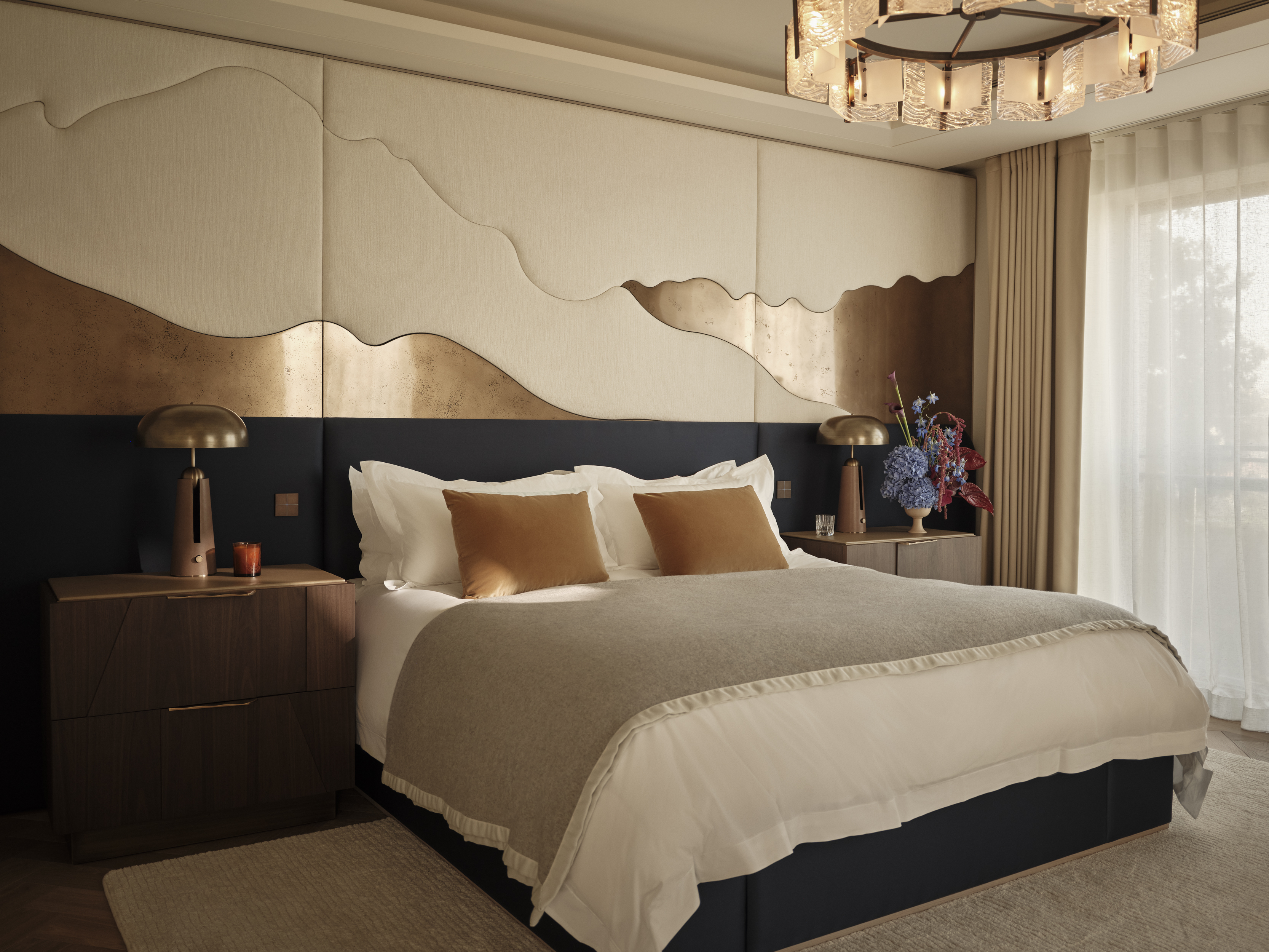 Bedroom with a navy upholstered headboard, cream bedding and ochre cushions beneath a sculptural wall panel with metallic accents.