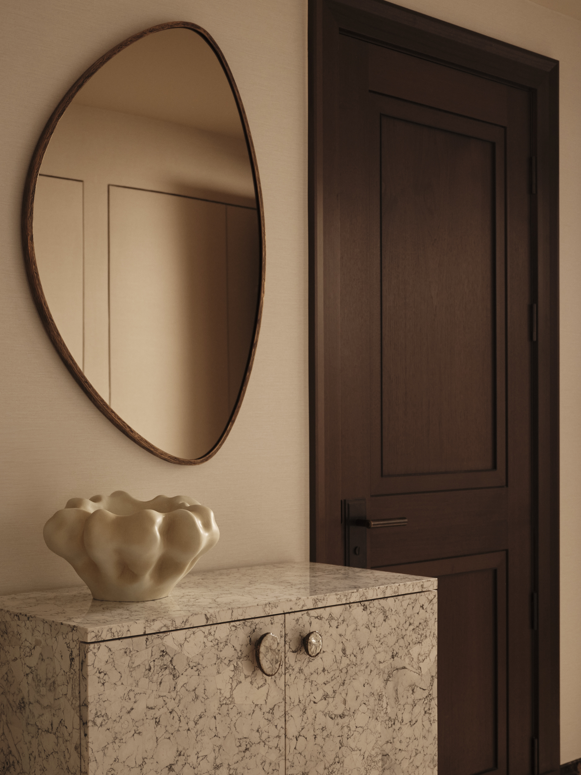 Irregular oval mirror above a textured pale cabinet, styled with a sculptural ceramic vessel beside a dark timber door.