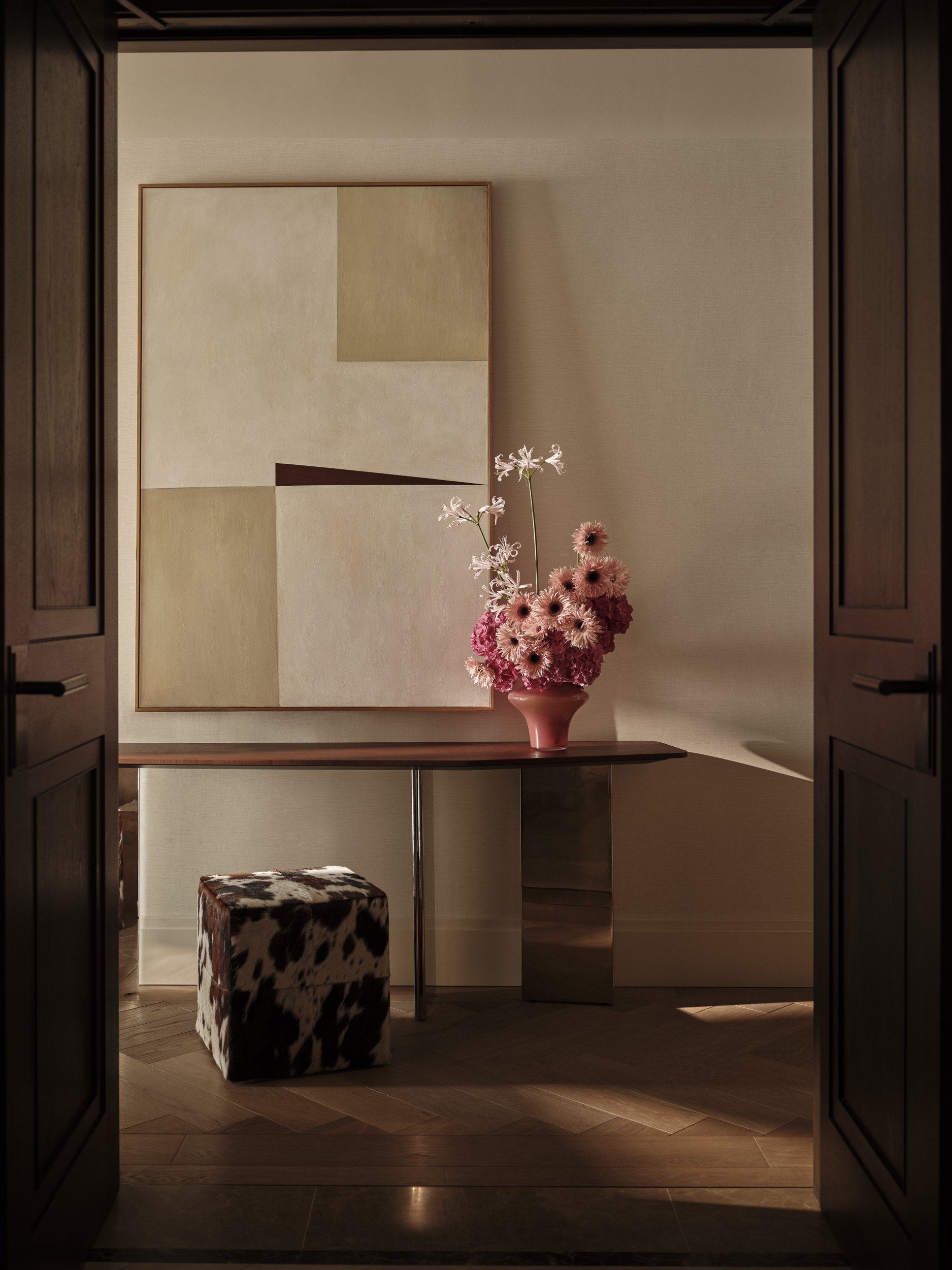 View through dark timber doors to a console table with pink flowers, abstract artwork and a patterned stool beneath.