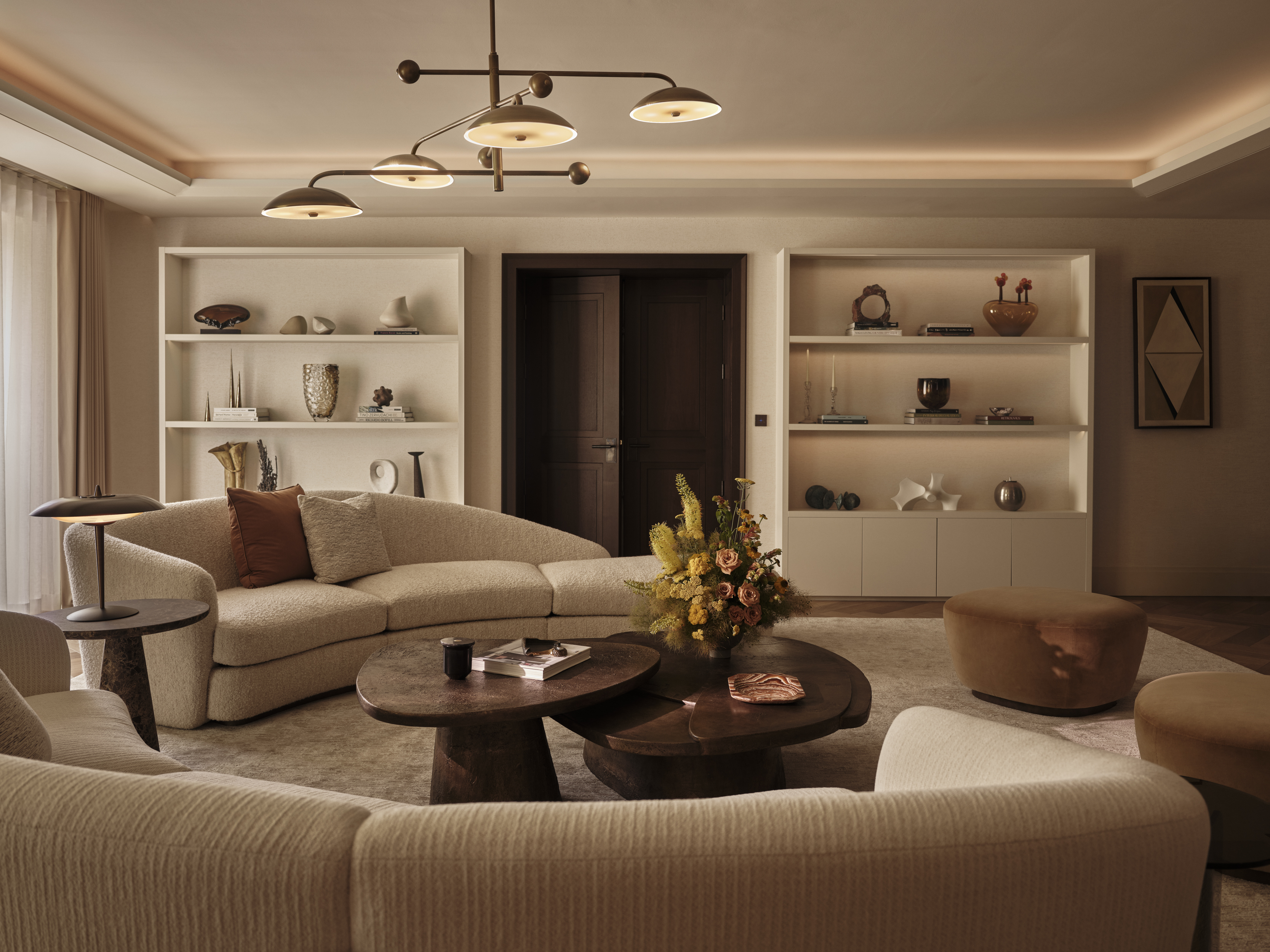 Cream sofas arranged around rounded timber coffee tables, with illuminated display shelving, sculptural objects and a branching ceiling light.