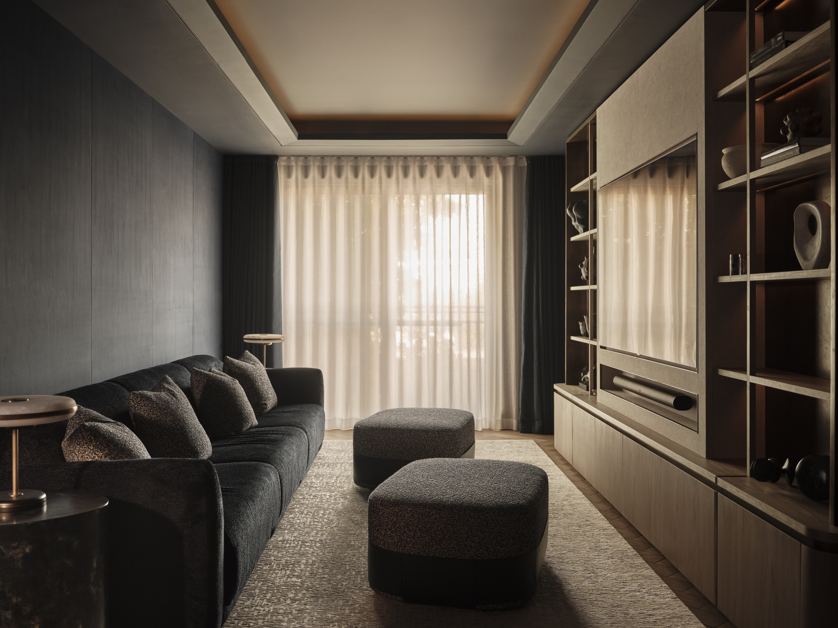 Dark upholstered sofa and two ottomans facing a television within timber cabinetry, with open shelving and full-length sheer curtains.
