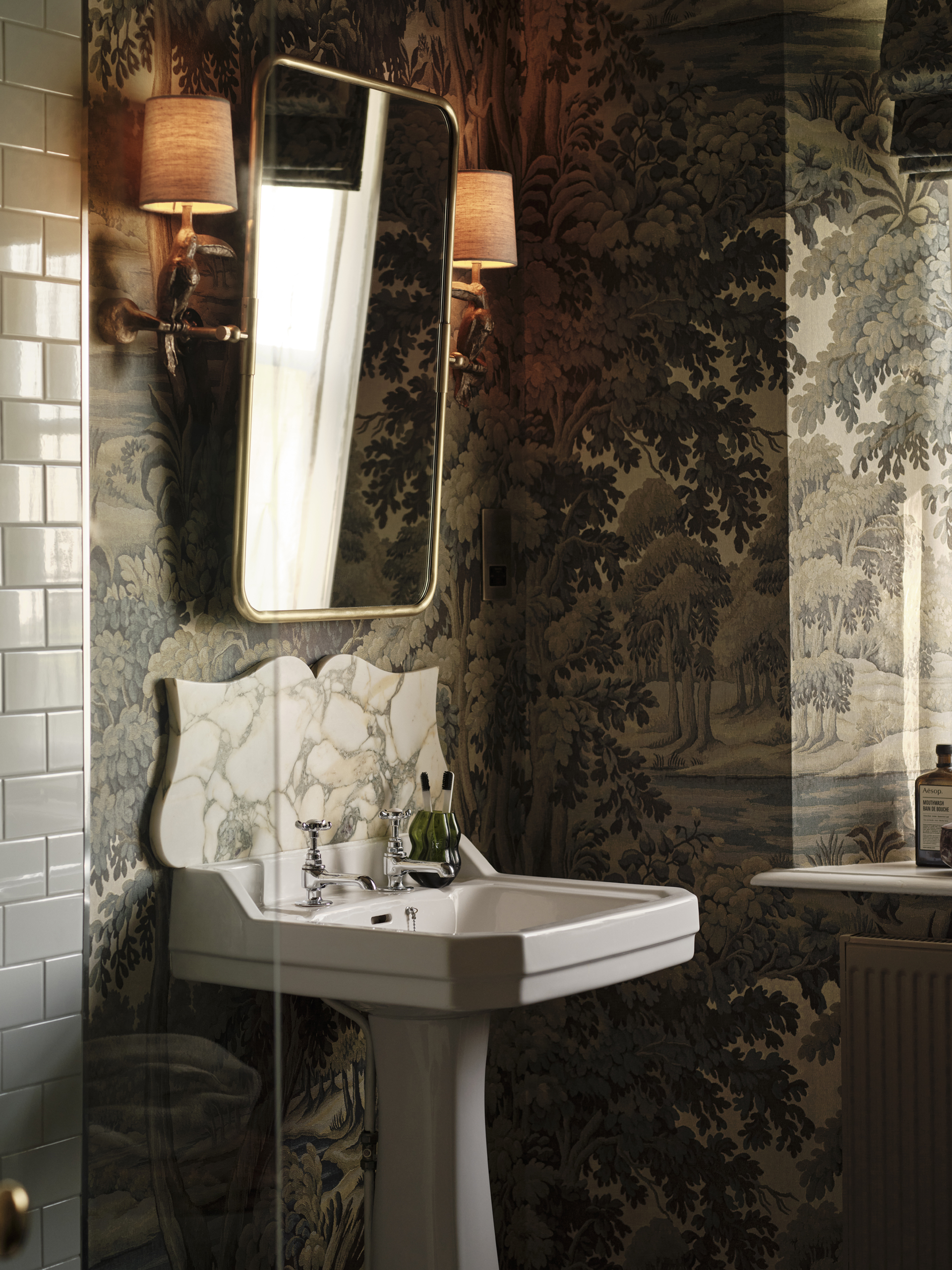 White pedestal basin beneath a brass-framed mirror and shaded wall lights, surrounded by botanical wallpaper and green tiles.