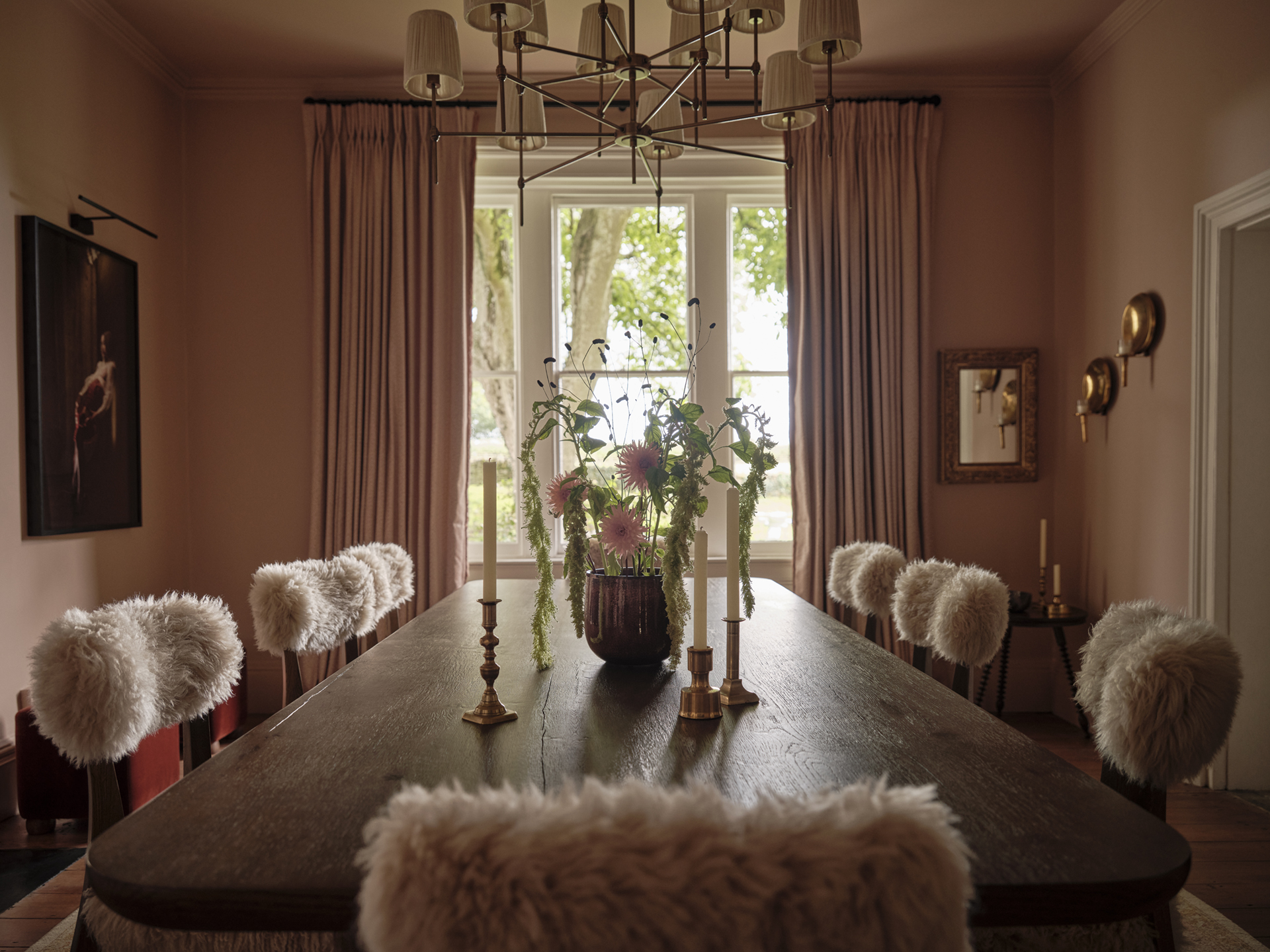 Long timber dining table with textured upholstered chairs, pink walls and full-length curtains framing a garden-facing window.