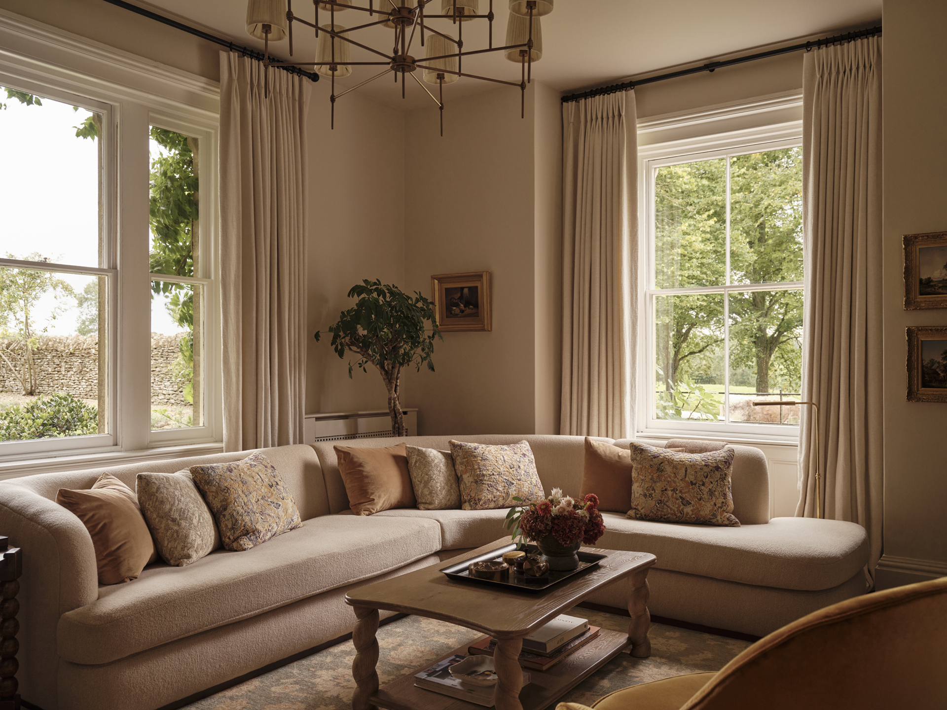 Curved cream sofa with patterned cushions arranged around a timber coffee table, beside tall windows overlooking the garden.