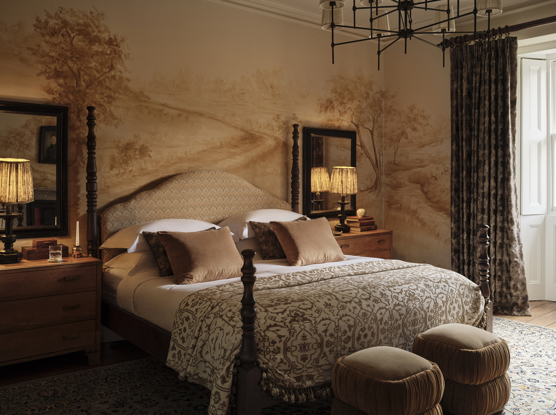 Four-poster bed against a softly coloured landscape mural, with timber bedside tables, patterned curtains and two upholstered stools.
