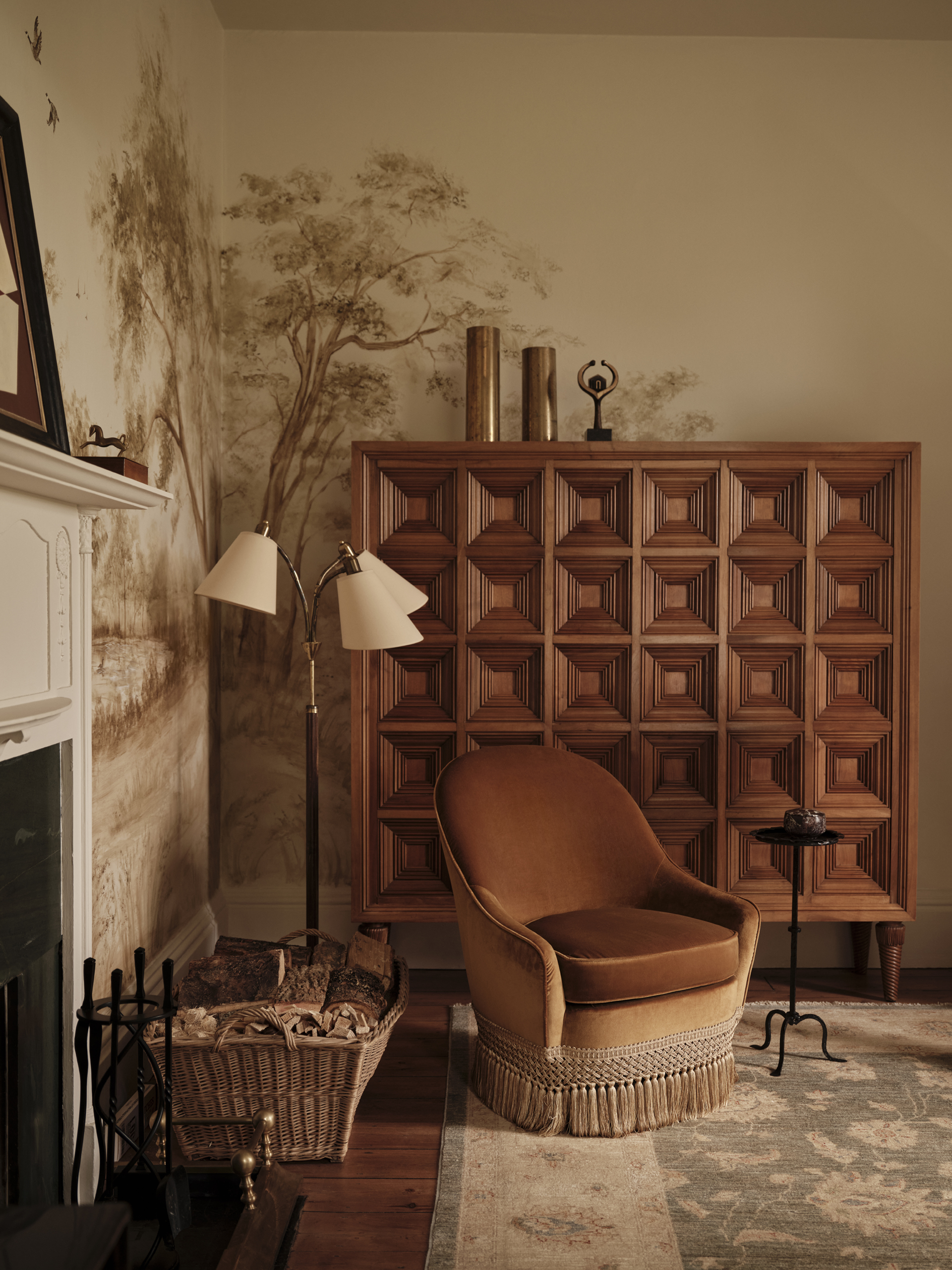 Ochre fringed armchair beside a fireplace, with a geometric timber drinks cabinet and a landscape mural behind.