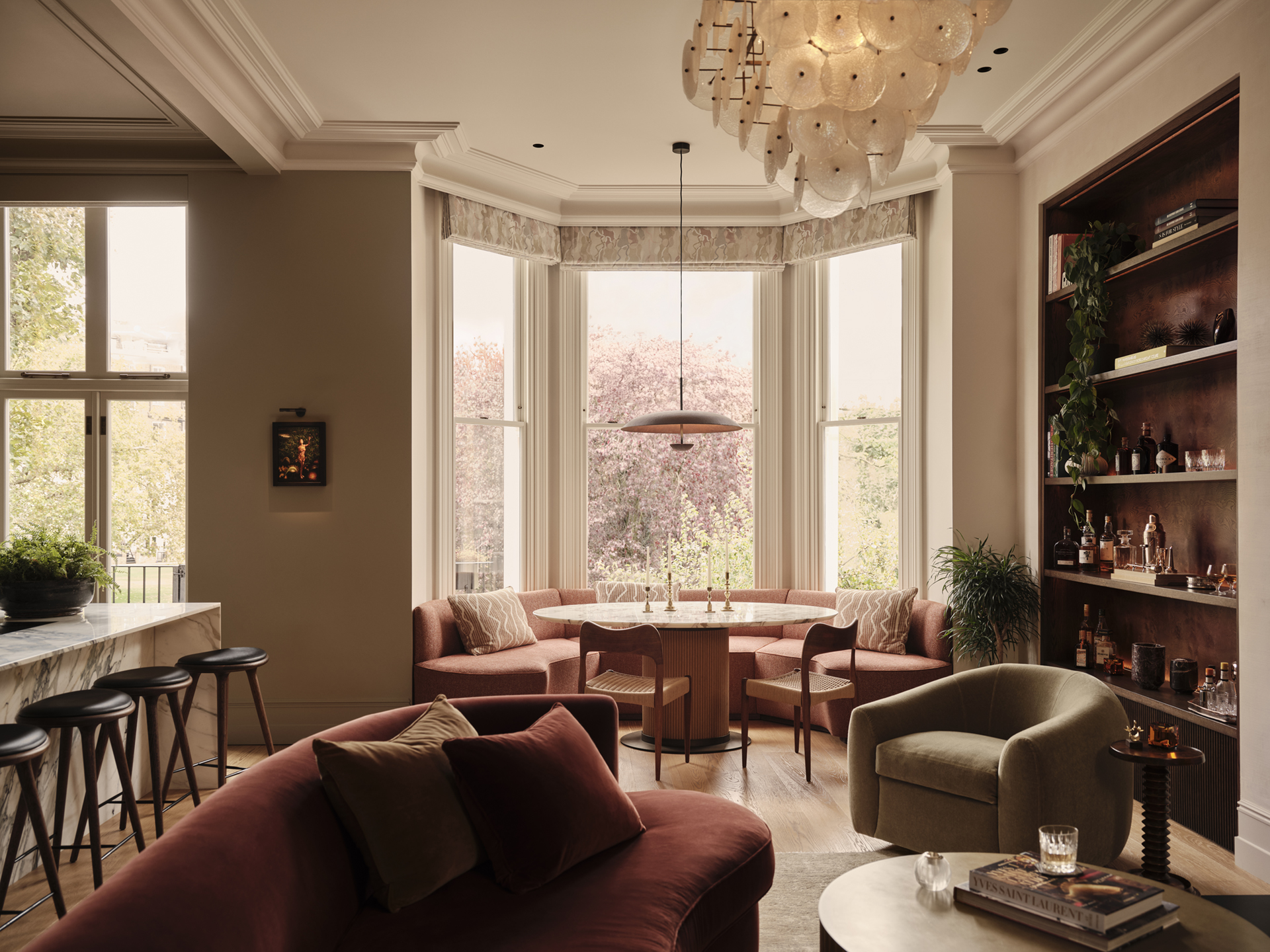 Dining table and upholstered seating within a tall bay window, overlooking an open-plan living area in a Maida Vale apartment by Designed by Woulfe.