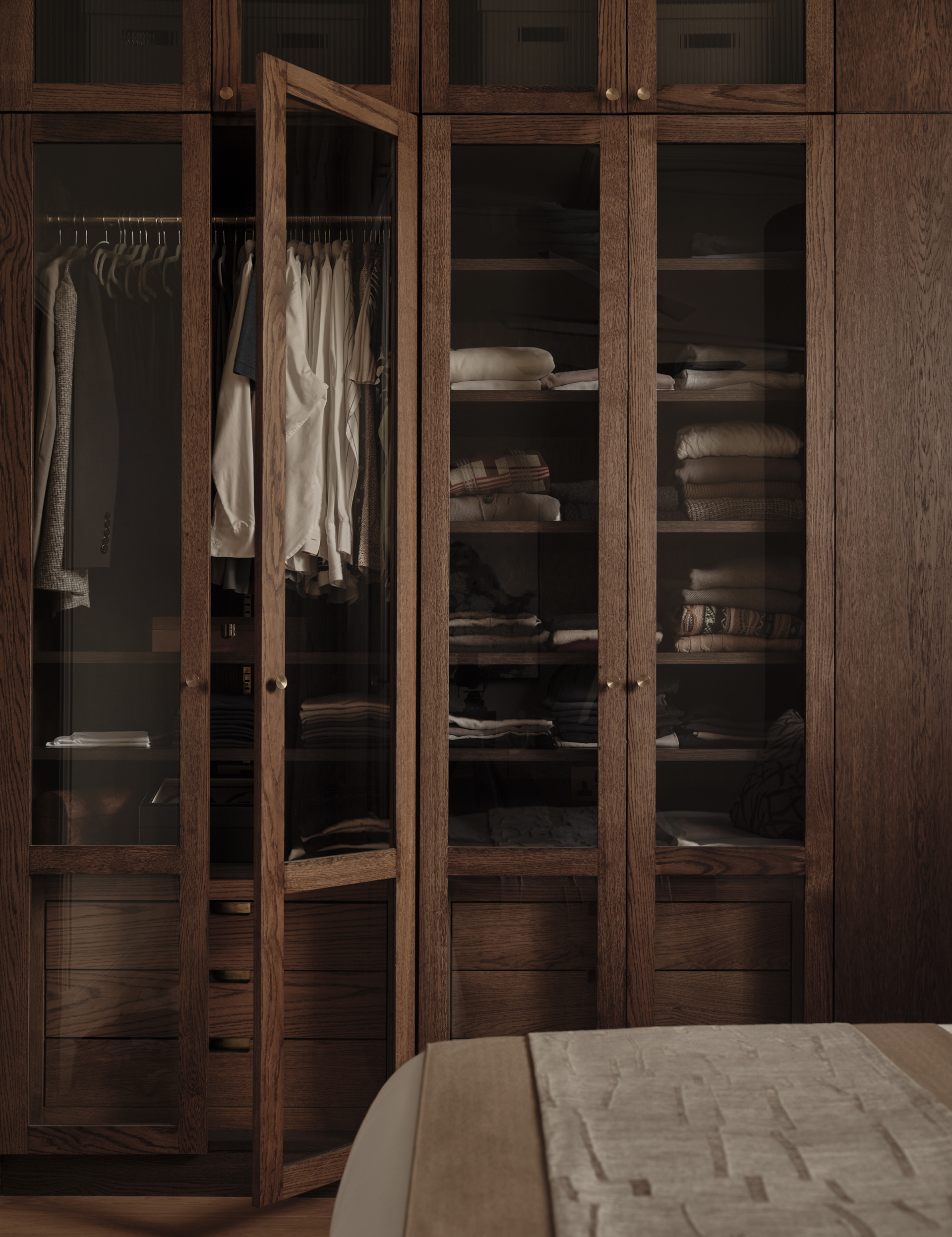 Bespoke dark wood wardrobe with glazed doors, integrated drawers and open hanging space in a bedroom by Designed by Woulfe.