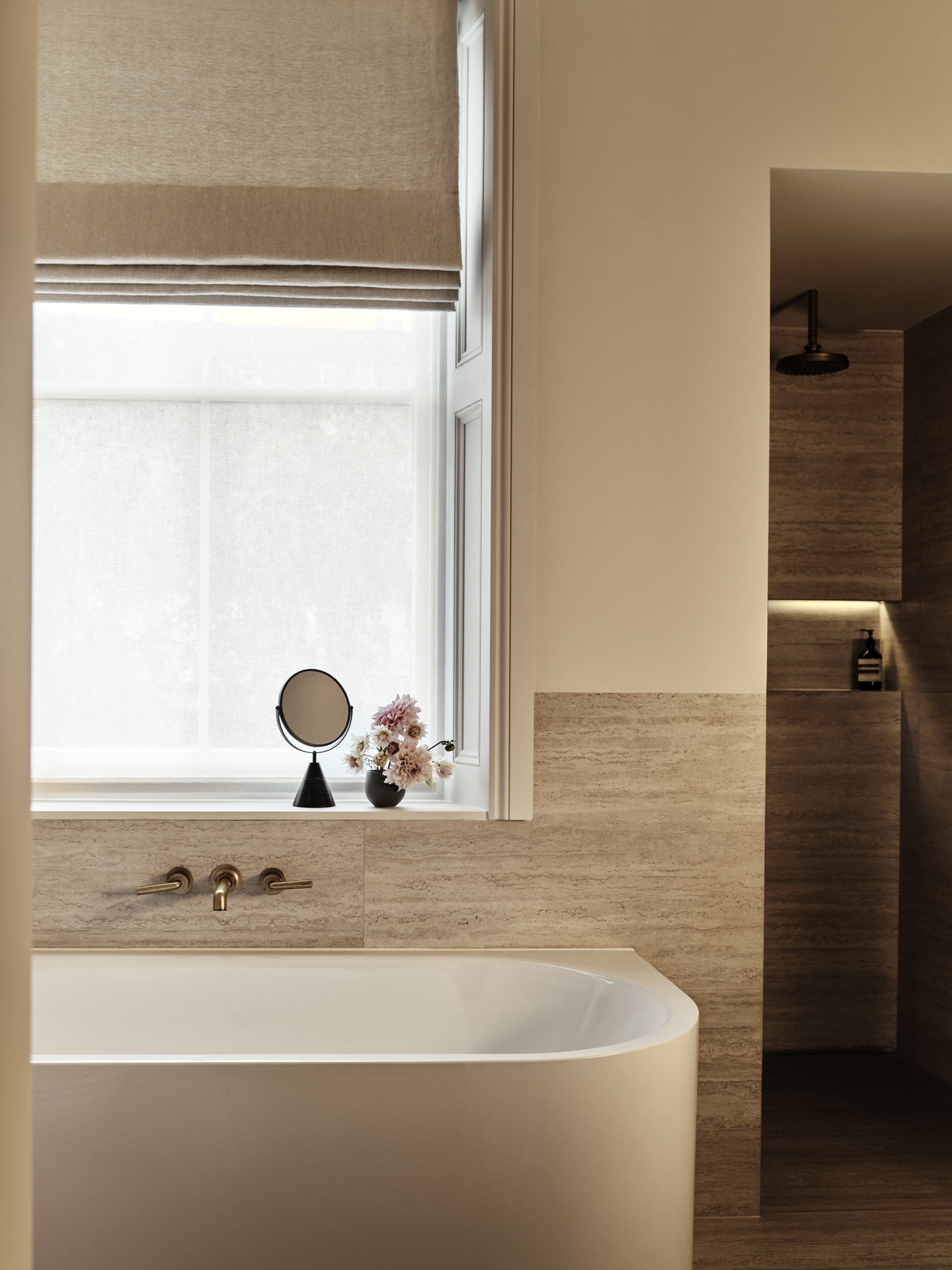 White freestanding bath beneath a window with a neutral Roman blind, beside warm-toned stone tiling and a walk-in shower.