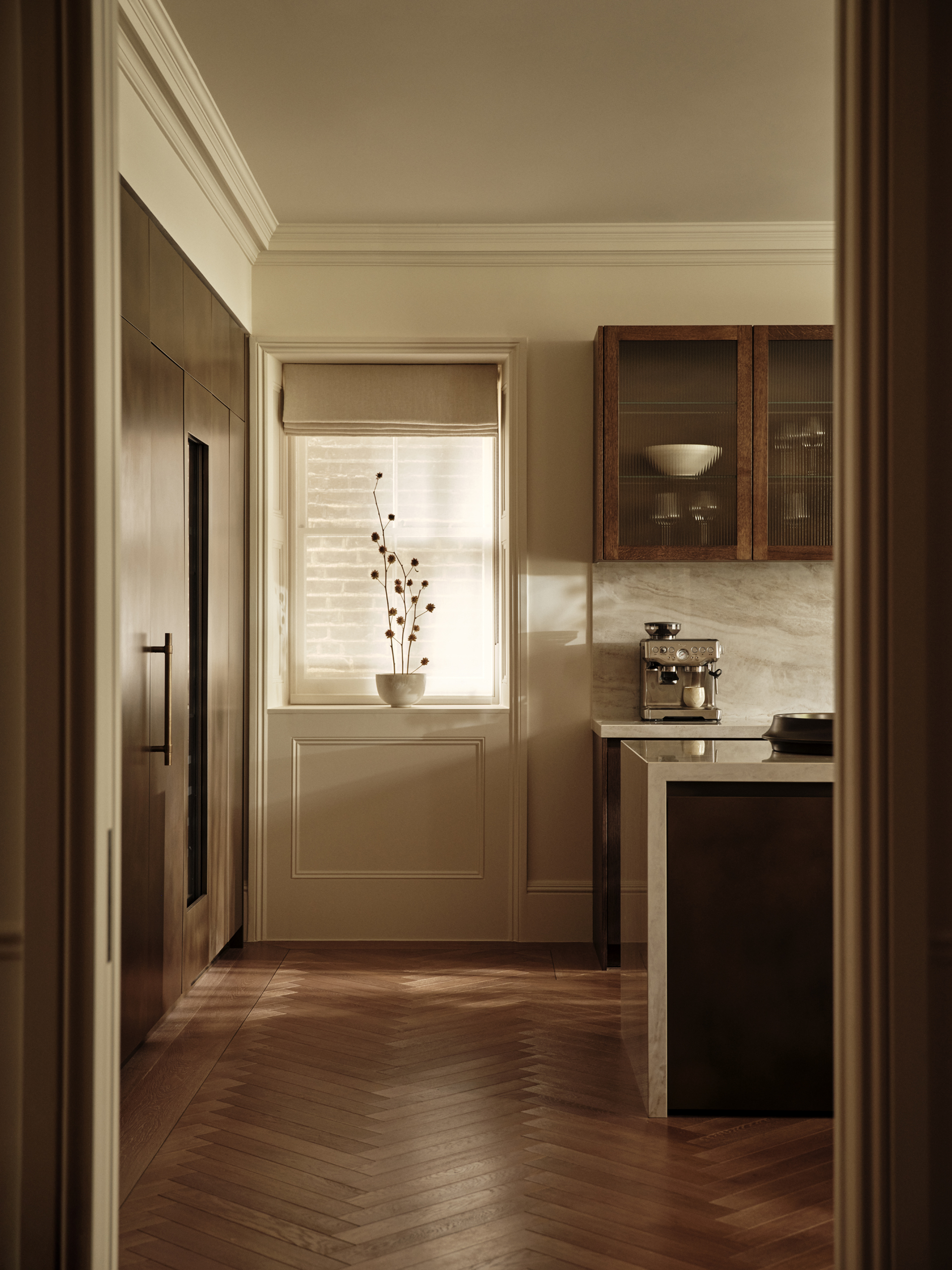 View through a timber doorway into a kitchen coffee corner, with glazed wall cabinetry, a Roman blind and herringbone flooring.