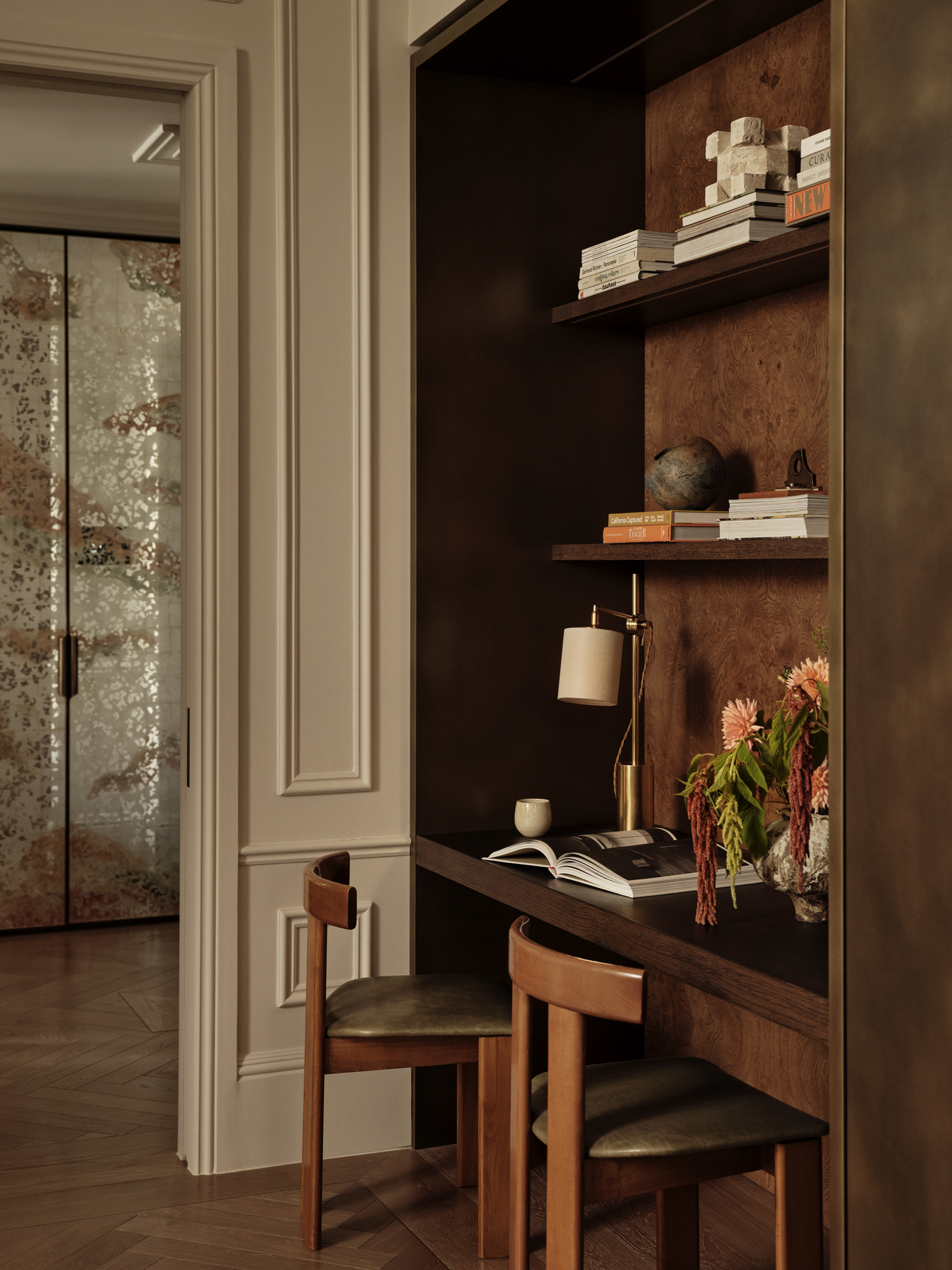 Recessed timber desk with two wooden chairs, open shelving, books and a small shaded lamp, set within a panelled wall.