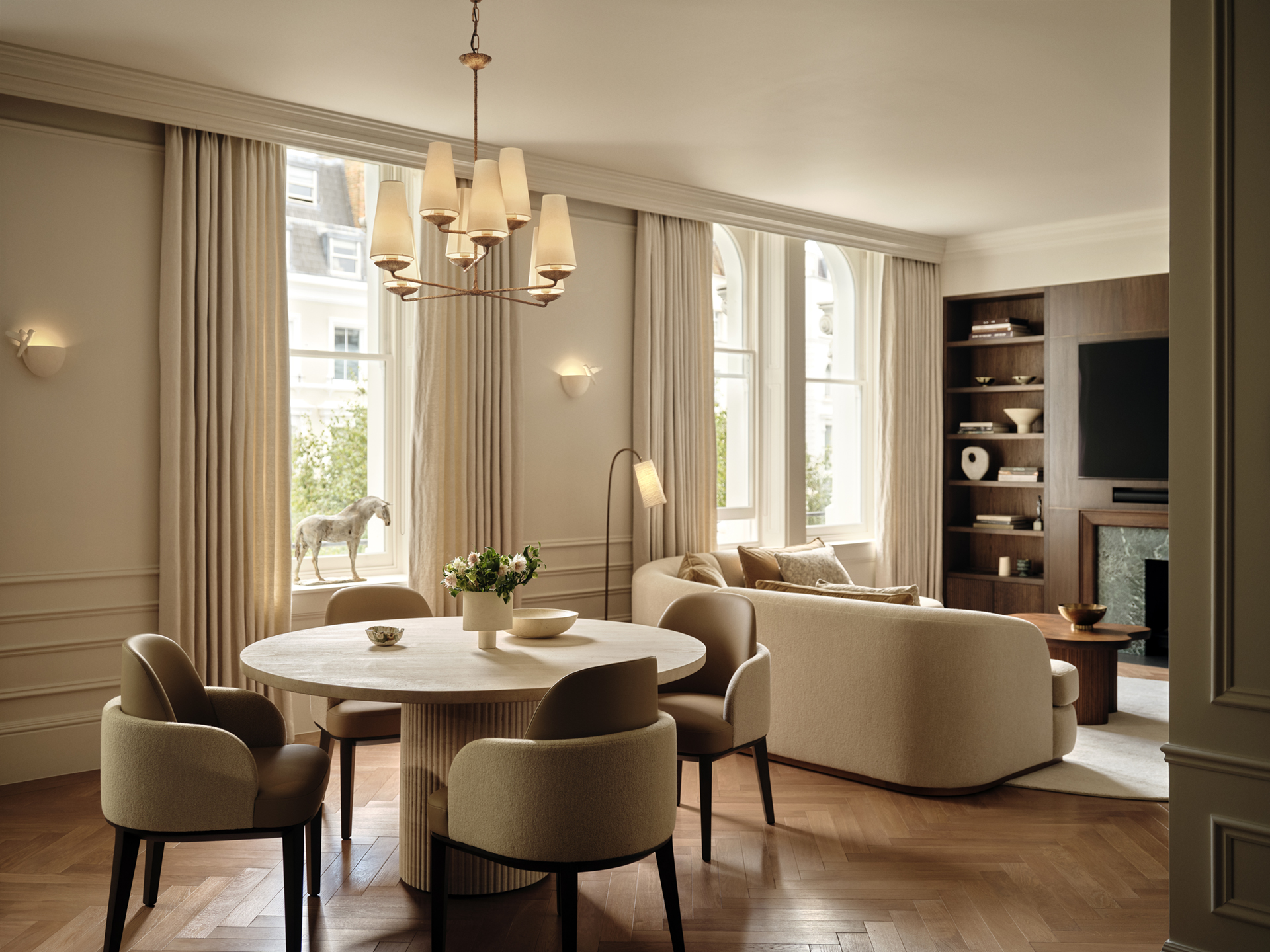 Round dining table with curved upholstered chairs opening onto a living area with cream seating, bespoke shelving and tall curtained windows.