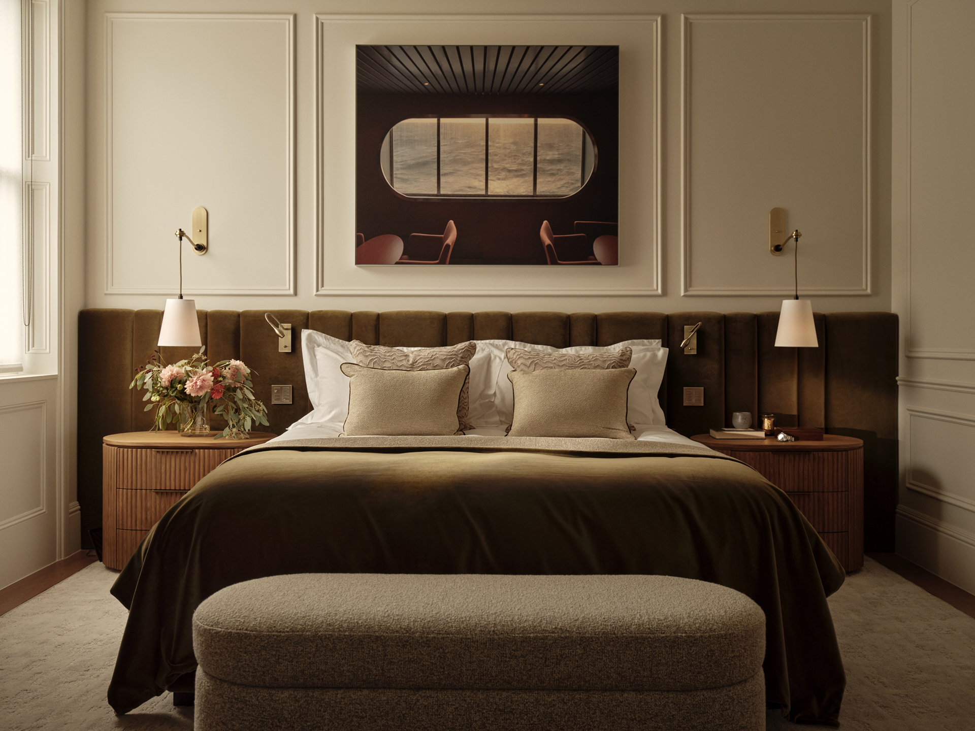 Bedroom with a dark timber headboard, cream pillows and a brown bedcover, with framed artwork above and an upholstered bench at the foot.