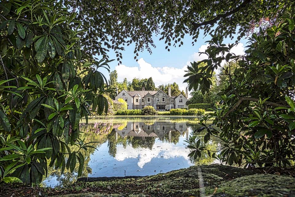 View of Moor Hall and Gardens in Aughton, Lancashire, framed by mature trees and reflected in the lake.