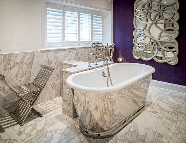 Luxury bathroom at Moor Hall, Aughton, featuring a freestanding marble-clad bath, marble flooring, white window shutters and a decorative mirror against a deep purple wall.