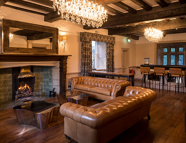 Restored lounge at Moor Hall, Aughton, featuring exposed timber beams, a traditional fireplace, leather Chesterfield seating and a crystal chandelier.