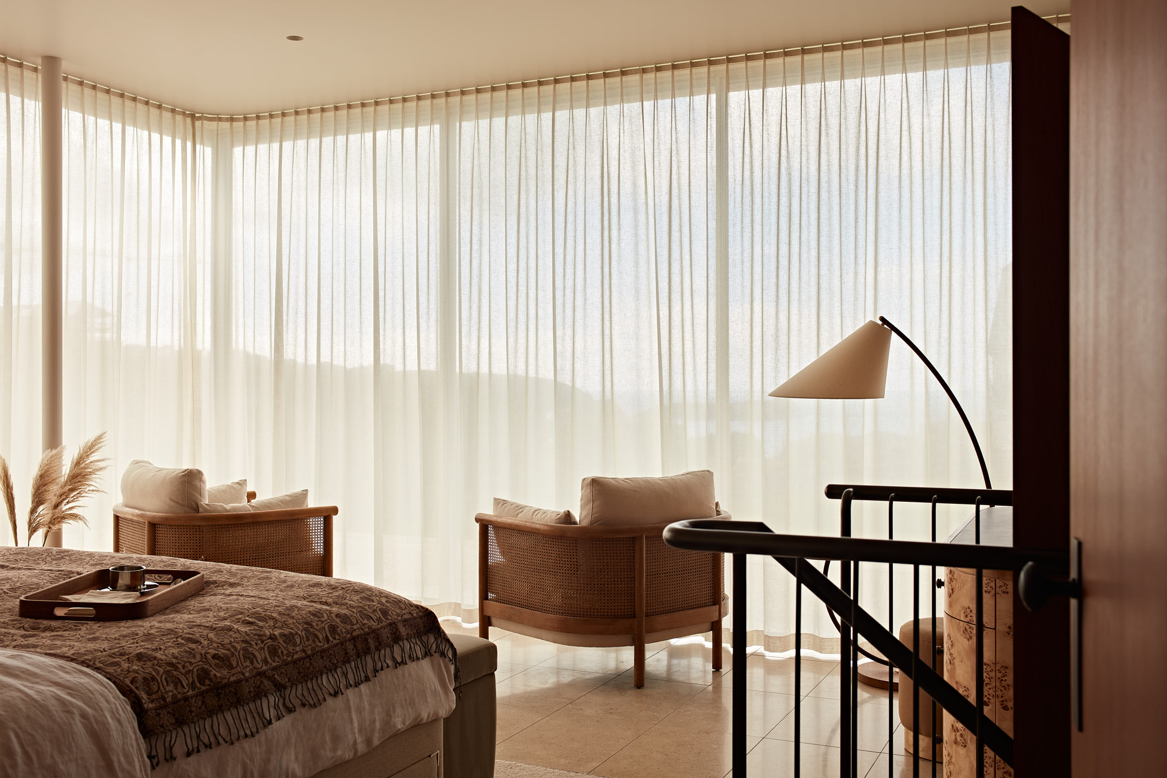 Bedroom with fully glazed corner windows, sheer drapes and modern cane arm chairs