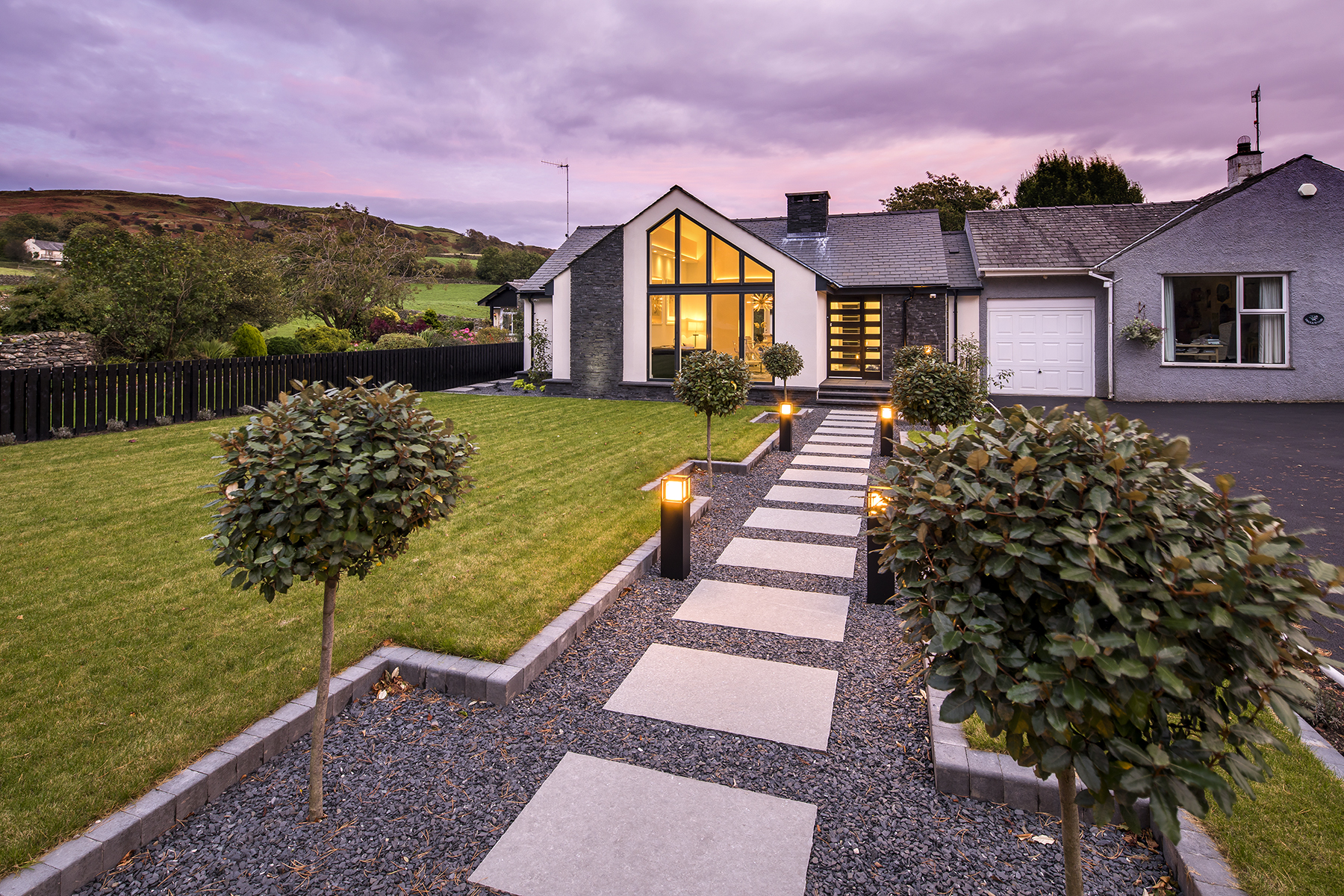 Transformed Lake District bungalow featuring a contemporary glazed entrance, natural stone detailing, landscaped gardens and illuminated pathway at dusk.